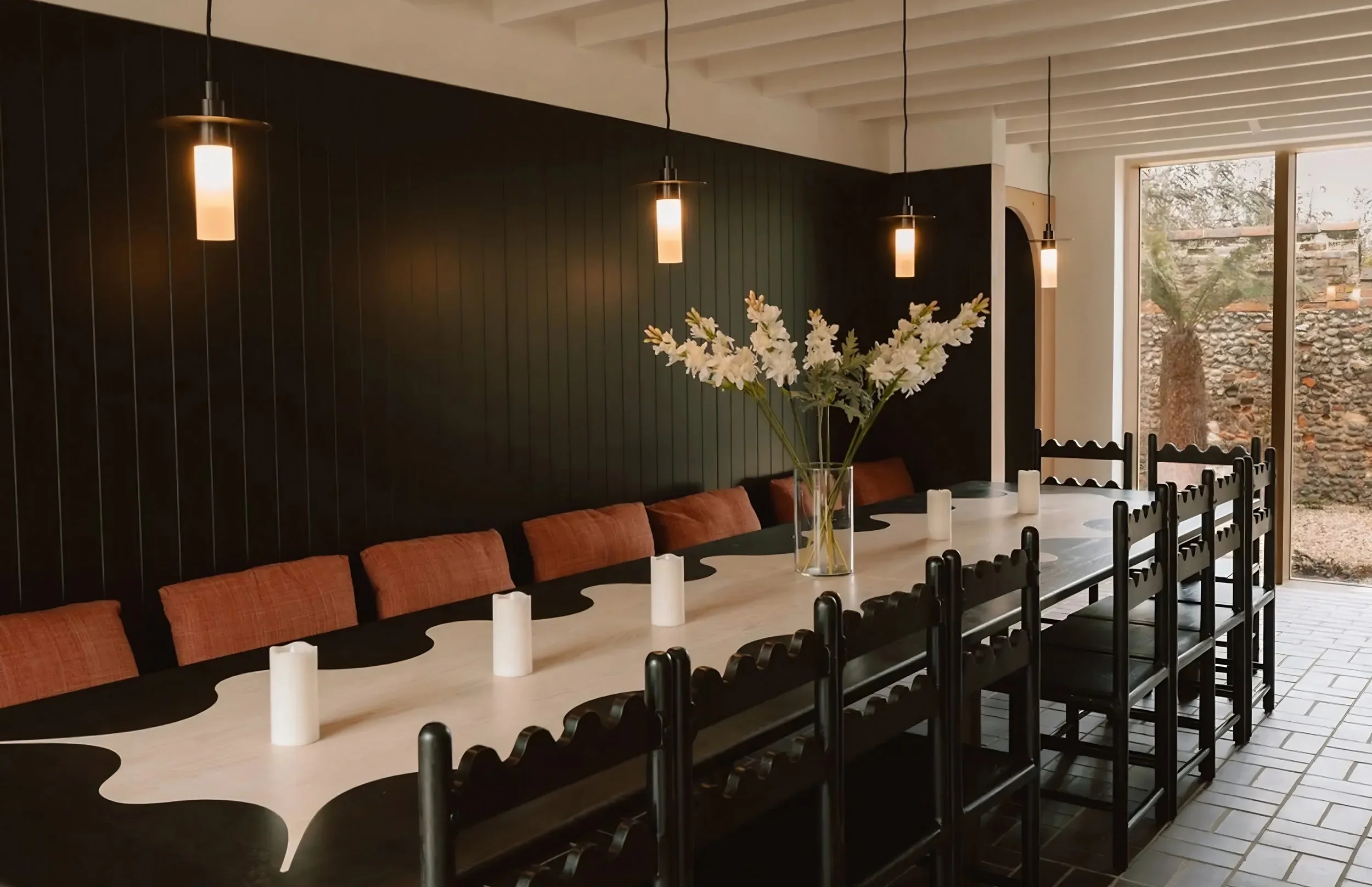 Dining room with black chairs around a light-colored table, a large glass vase with white flowers as a centerpiece, and black pendant lights hanging from a white ceiling. Large windows reveal an outdoor area with a brick wall and trees.