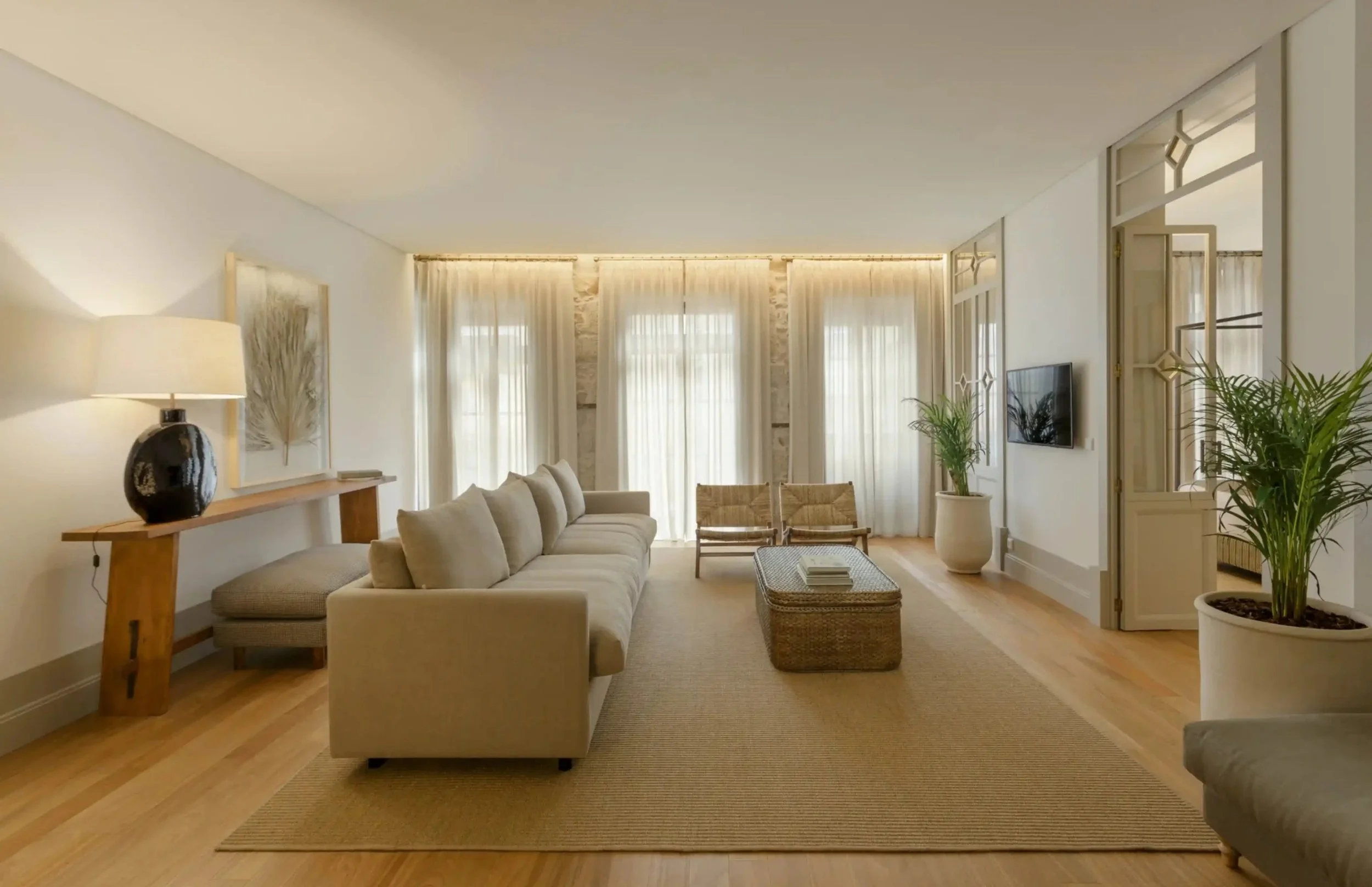 A living room with a beige sofa, wicker coffee table, large potted plants, cream curtains, and a wall-mounted TV.