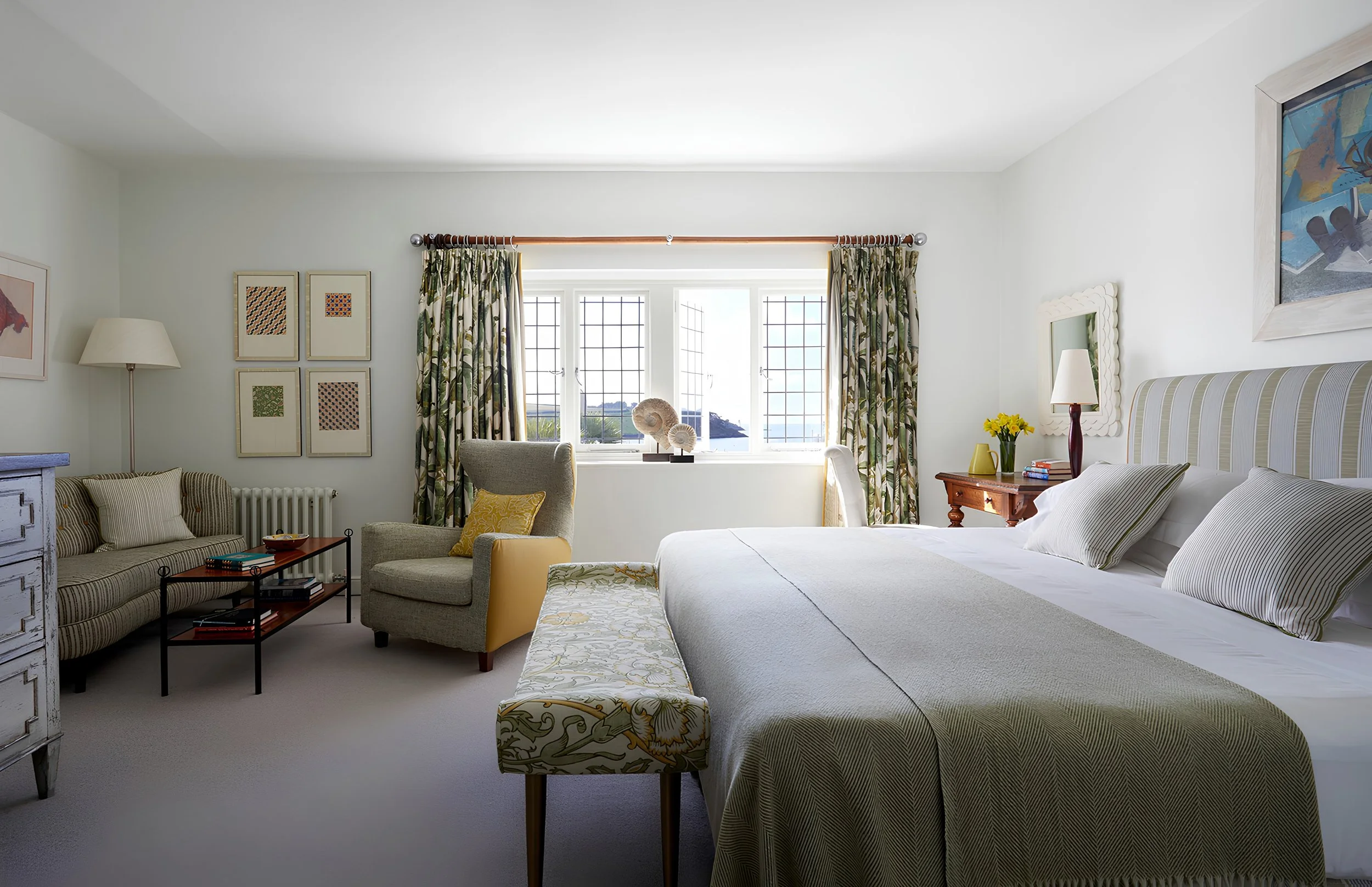 A bright and airy bedroom with white walls and a large window overlooking a scenic view. The room features a bed with white linens, a wooden side table with a lamp and yellow flowers, a cozy armchair with a yellow pillow, framed artwork on the walls,