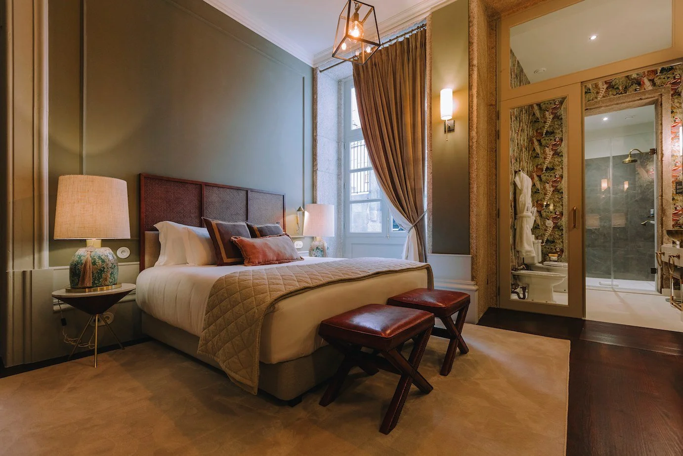 A cozy hotel bedroom with a large bed, bedside lamps, decorative pillows, a window with brown curtains, and a bathroom with a shower visible through a glass door.