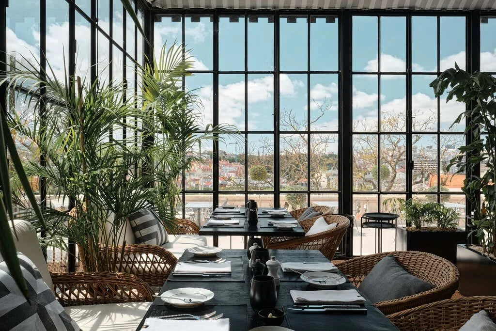 A sunny indoor dining area with large floor-to-ceiling windows, wooden tables set with plates, napkins, and utensils, wicker chairs, and various indoor plants, overlooking a cityscape with trees and buildings.