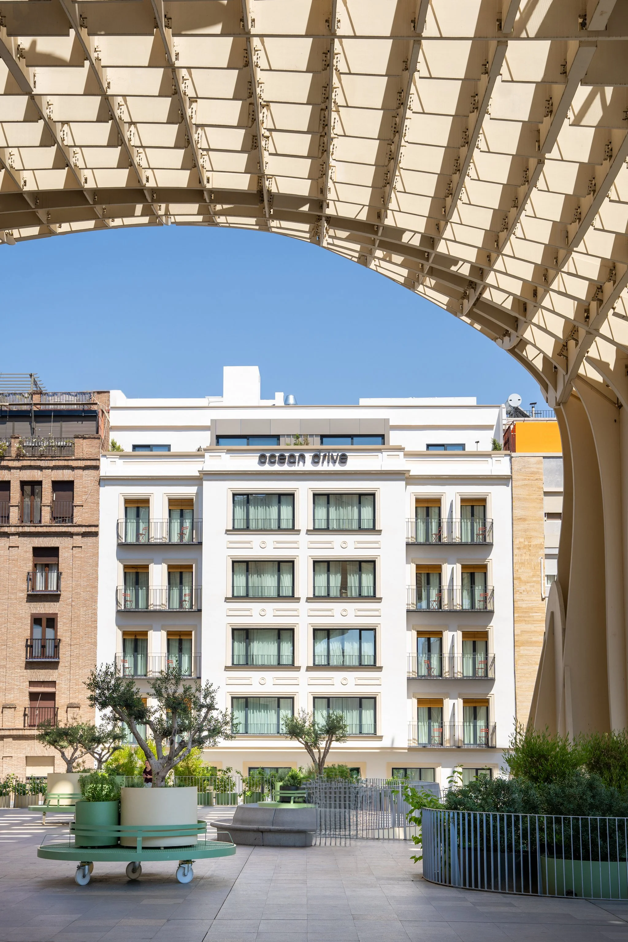 View of a modern city street with a white building labeled "ocean drive" and greenery in the foreground, seen through a large, arched metal structure overhead.
