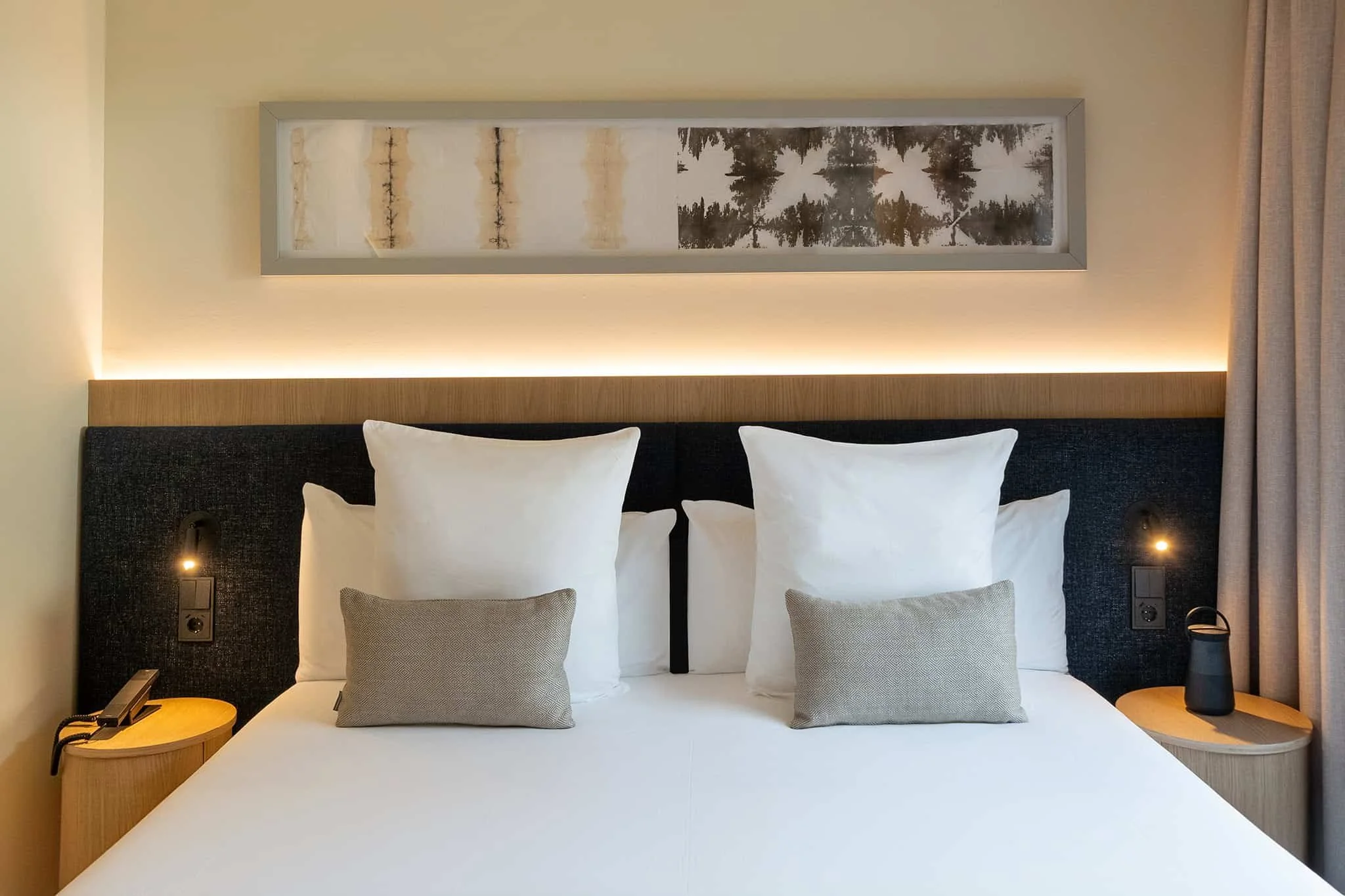 A neatly made hotel bed with white sheets, multiple white pillows, and two gray decorative pillows. Behind the bed is a black upholstered headboard, framed artwork of black and white trees, beige walls, and a small round wooden nightstand with a black lamp and a black water bottle on it.
