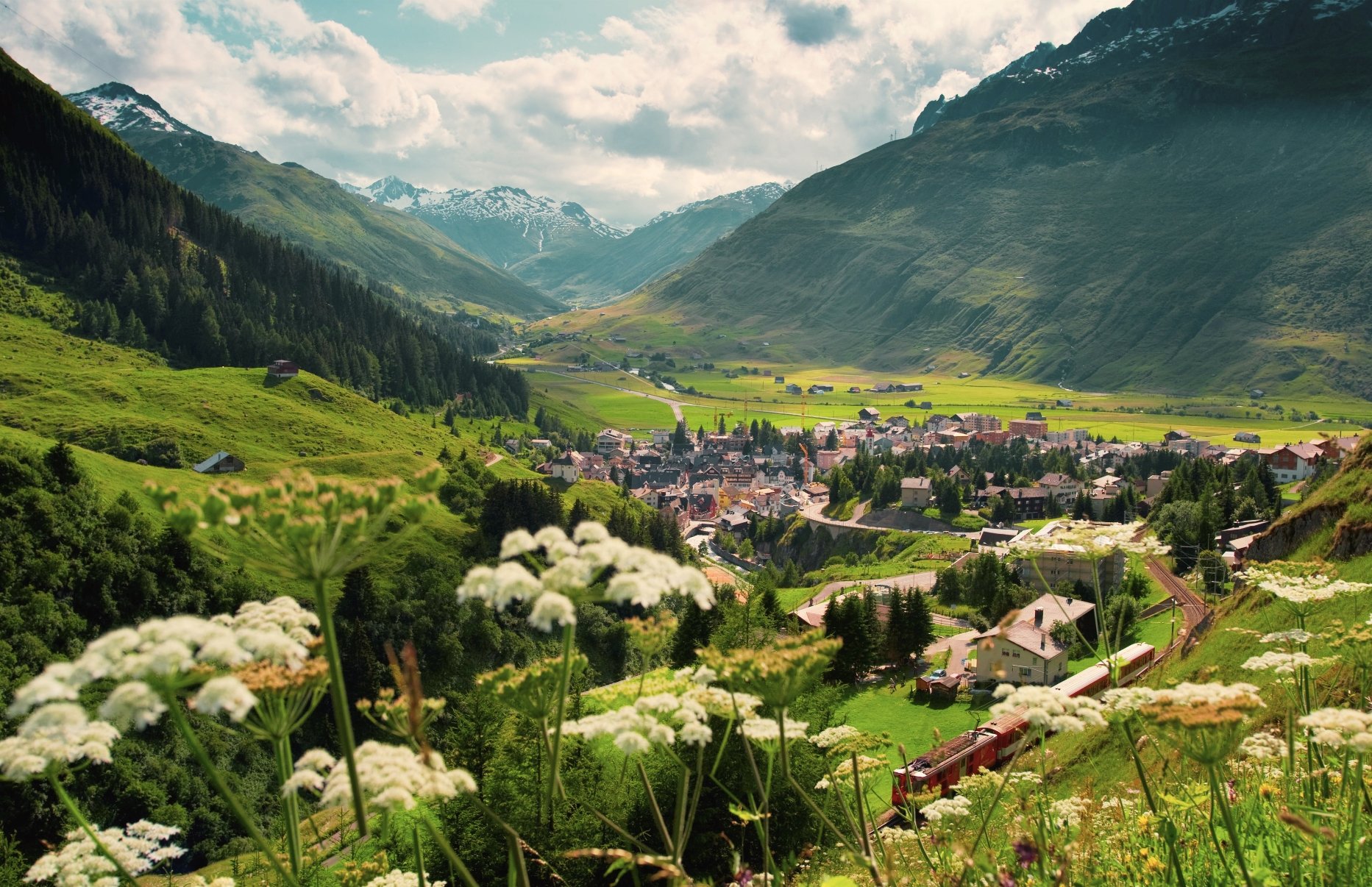 A scenic view of a small town in a valley, surrounded by green hills and snow-capped mountains, with flowers in the foreground and partly cloudy sky overhead.