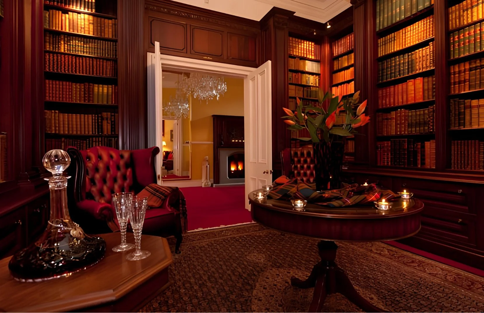 An elegant room with dark wood bookshelves filled with books, a red velvet armchair, a table with glasses and a decanter, a round table with candles, a large flower arrangement, and a fireplace in the background.