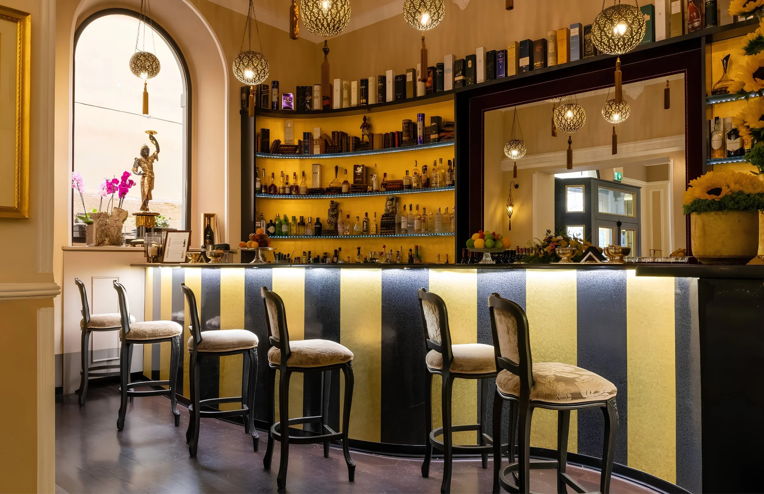 Interior of a bar with yellow and black striped counter, high chairs, and shelves filled with liquor bottles. Decorative hanging lamps and a large window with a statue and pink flowers on the sill.