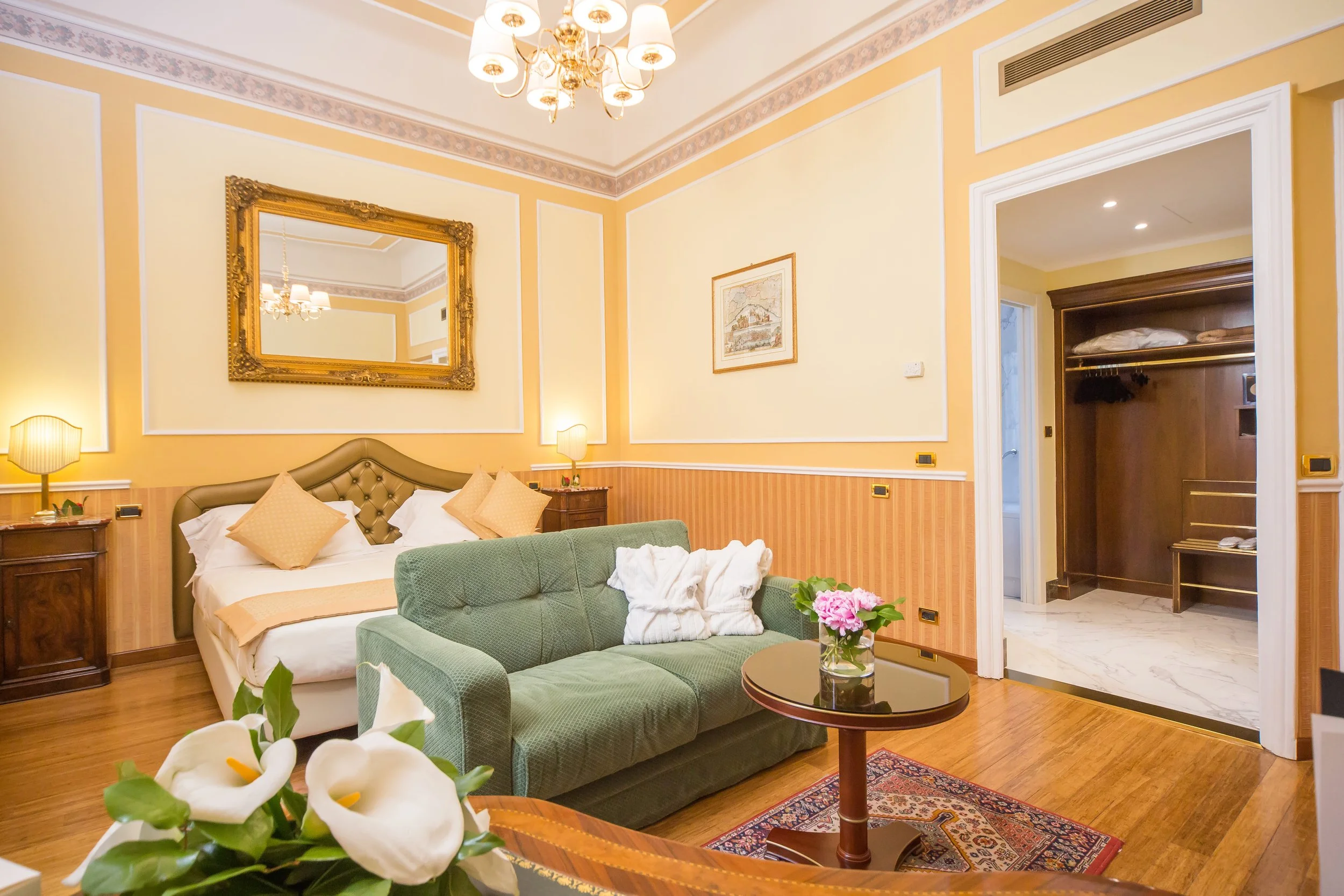 A hotel room with yellow walls, a bed, a green armchair with pillows, a wooden bedside table, a large gold-framed mirror, decorative painting, a chandelier, a doorway leading to a closet with shelves, and a small round table with a flower arrangement.