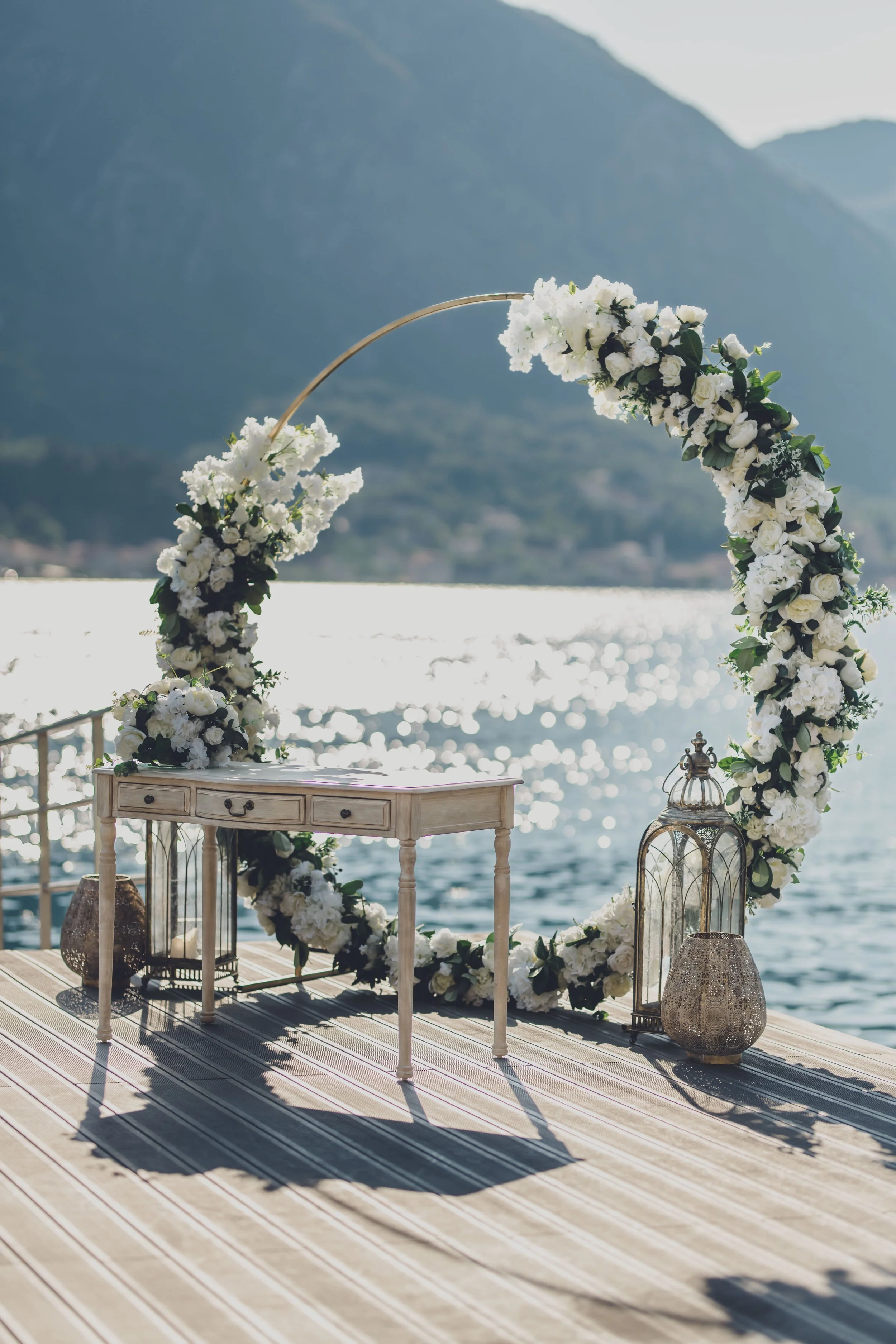 A wedding arch decorated with white flowers and greenery, positioned on a wooden deck by a lake with mountains in the background, during daytime.
