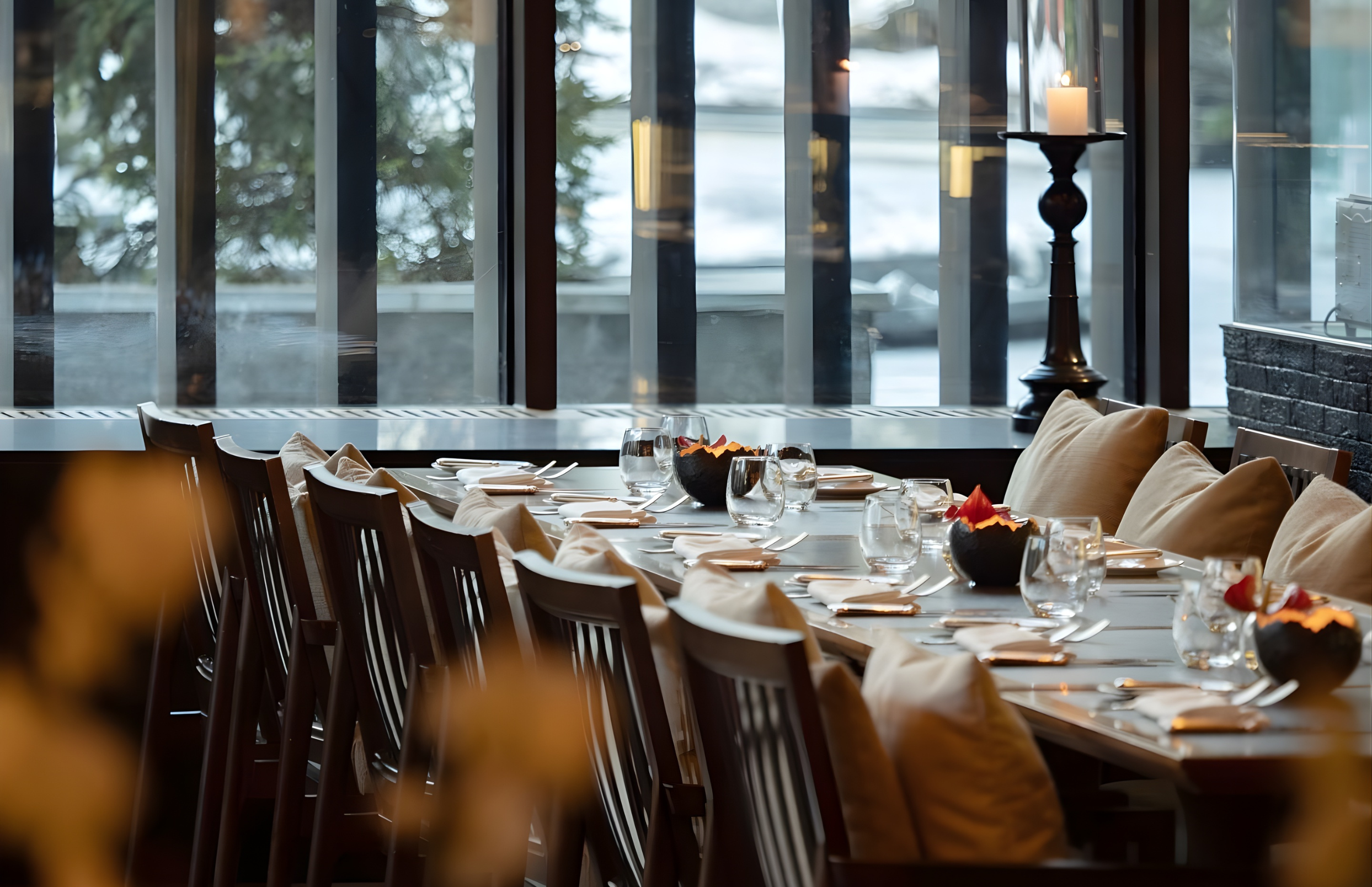 A dining table set with white napkins, glasses, and desserts inside a restaurant near large windows with views of trees and snow outside.