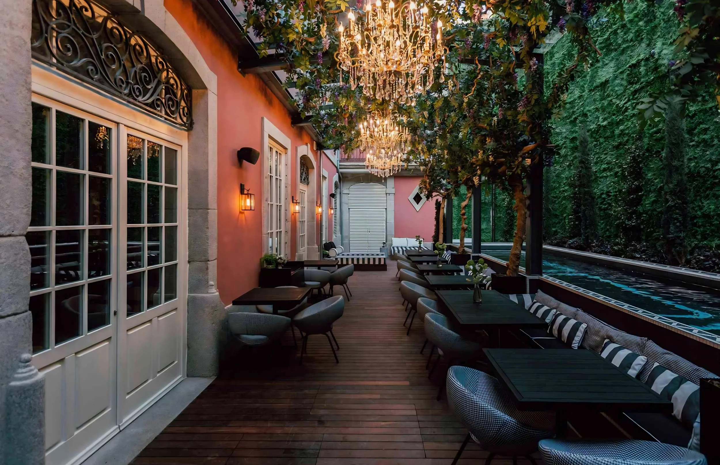 A beautifully decorated outdoor patio with chandeliers hanging from a ceiling covered with green foliage. The patio has wooden flooring, stylish chairs, and tables with small flower arrangements. Pink walls with windows and sconce lighting add to the