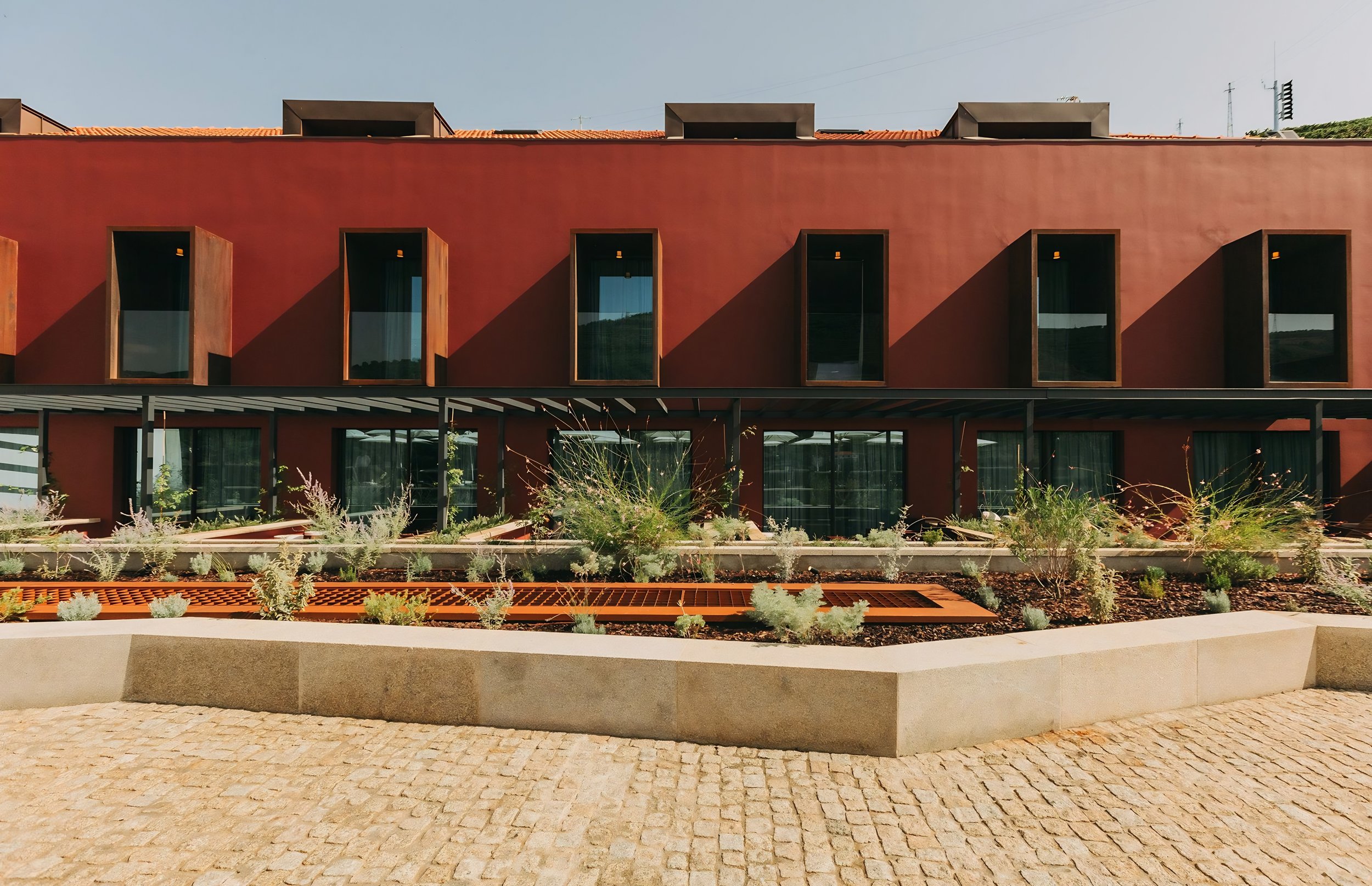 Modern building with red exterior, multiple rectangular windows with wooden frames, and a garden area with plants in front. Ground paved with cobblestones.