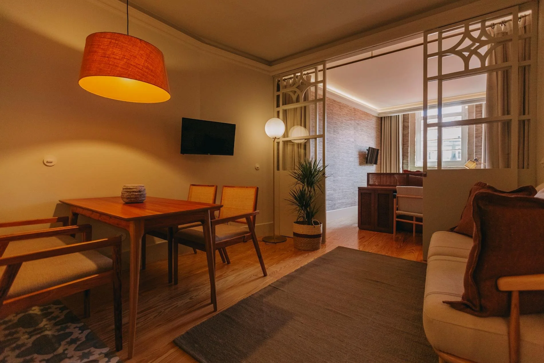 Living room with a wooden dining table and chairs, a beige sofa with brown pillows, a potted plant, wall-mounted TVs, and warm lighting from a hanging lampshade and floor lamps, with large windows.