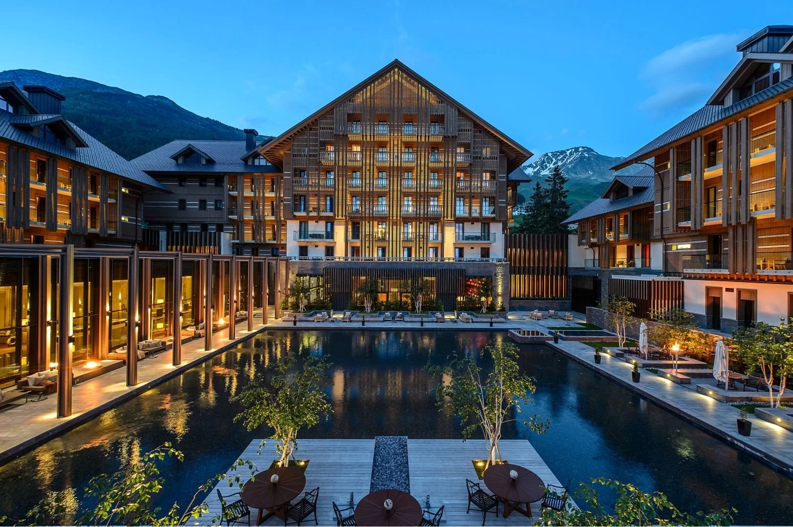 A modern hotel complex at dusk with an illuminated pool in the foreground, surrounded by wooden and glass structures, with mountains in the background.