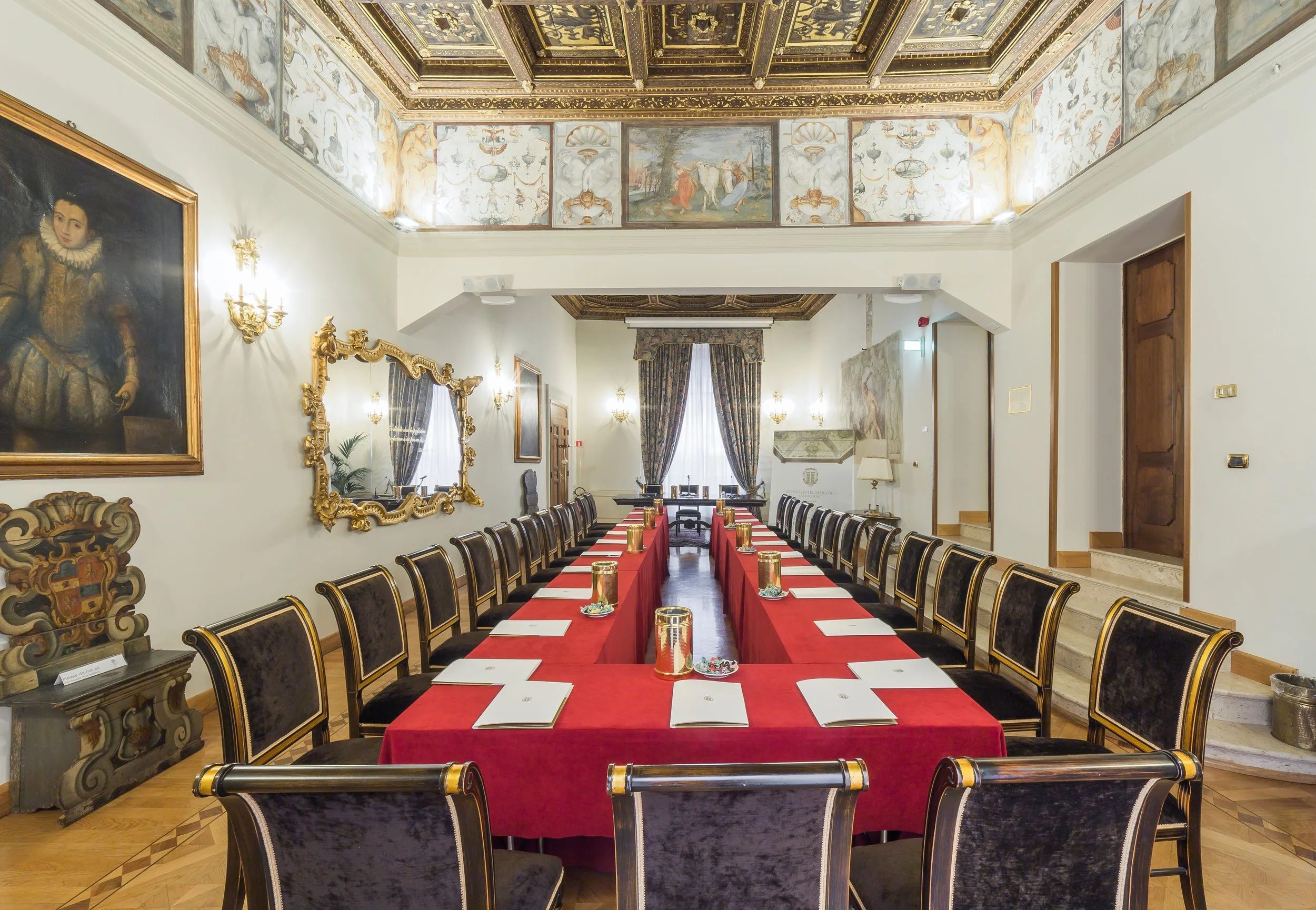 A large conference room with a long table covered with a red cloth and set with white documents and gold containers. The room has elegant, ornate decor with a gilded mirror, paintings, and a ceiling with intricate gold detailing and artwork. Tall windows with heavy curtains are at the far end, and chairs with velvet upholstery are arranged around the table.