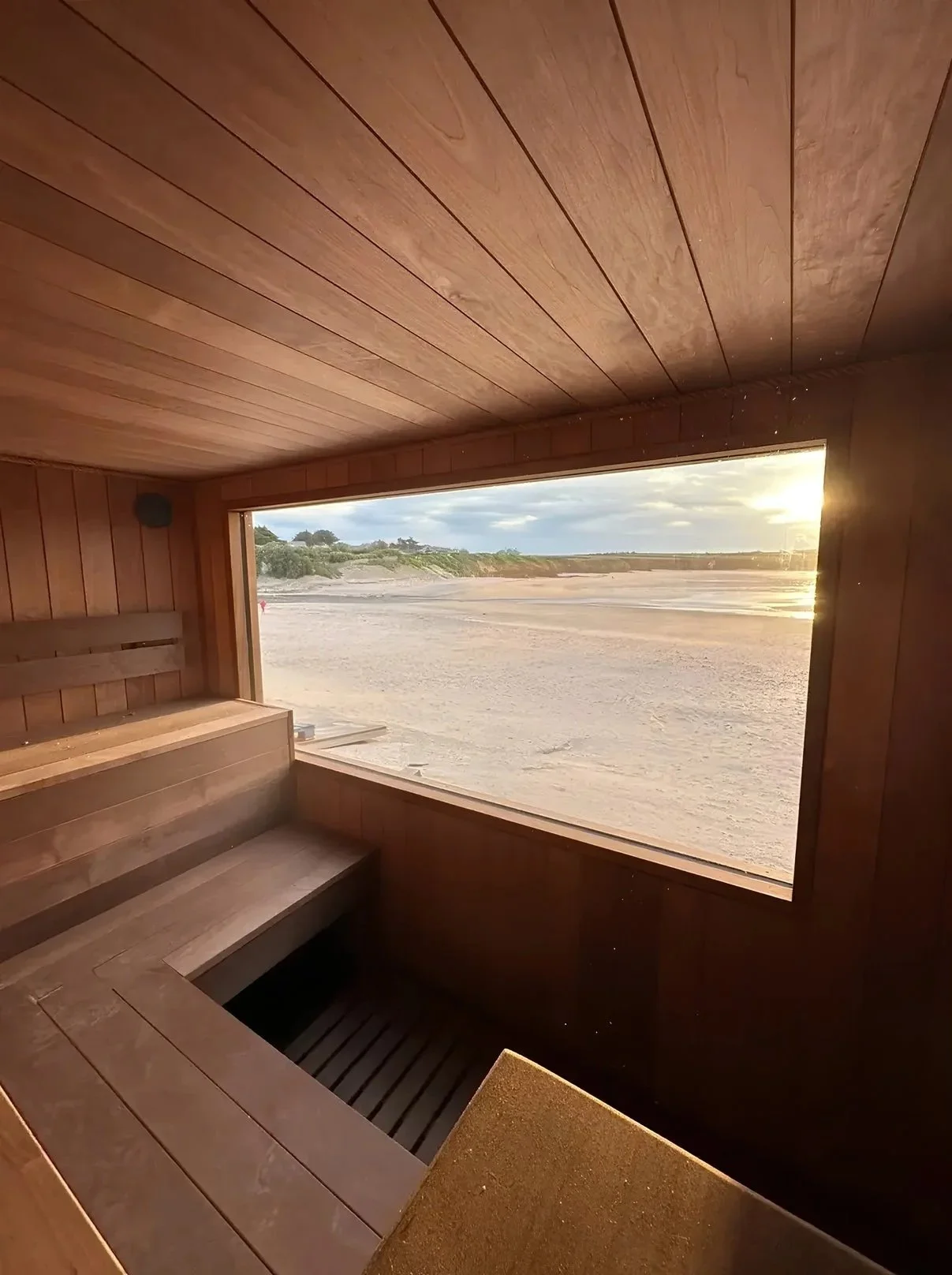 Wooden sauna room with a large window overlooking a sandy beach and ocean at sunset.