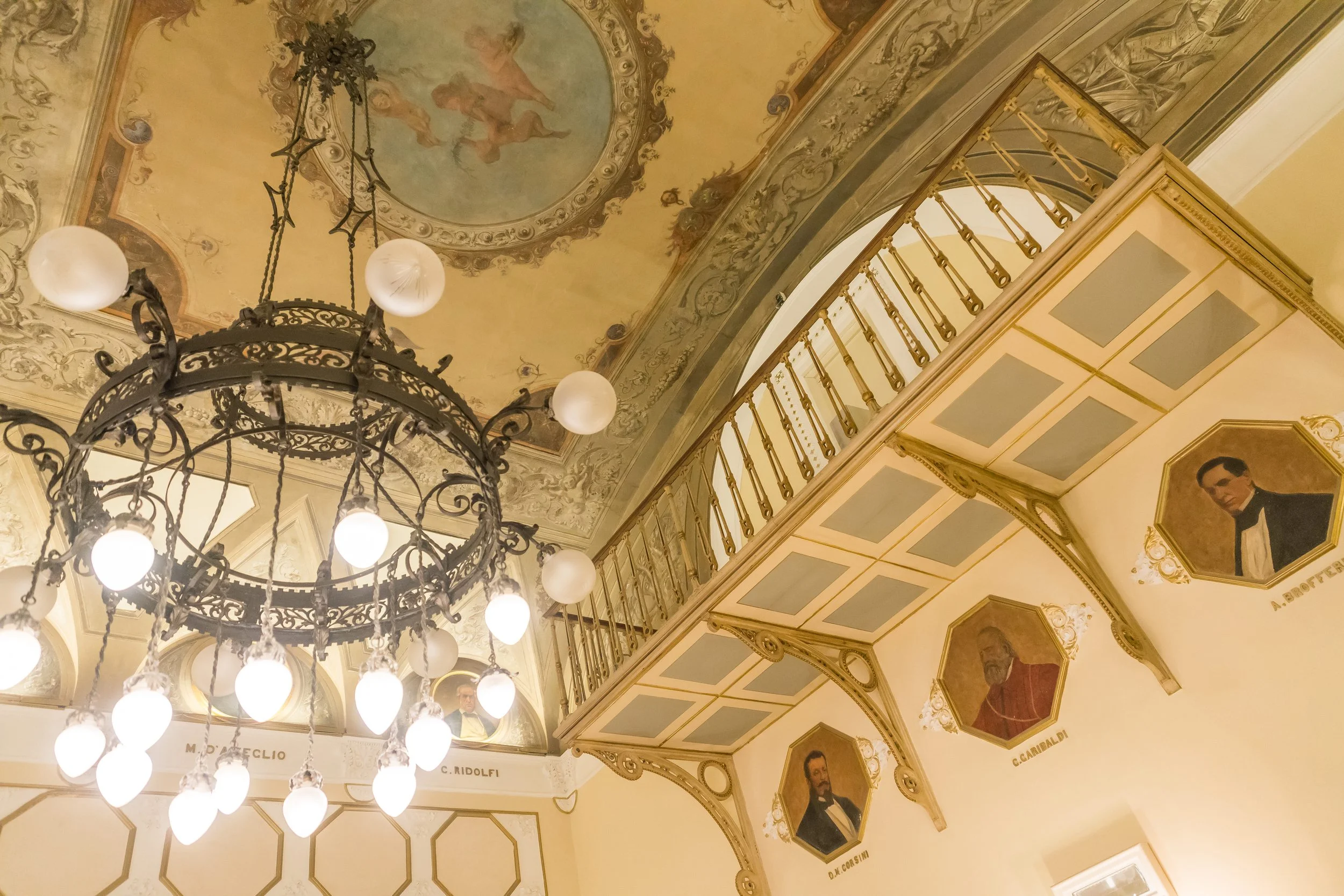 Elegant ornate ceiling with a painted cherub or angel surrounded by decorative moldings, a large black wrought iron chandelier with spherical frosted glass bulbs, and portraits of distinguished individuals framed on the wall below, in a luxurious int