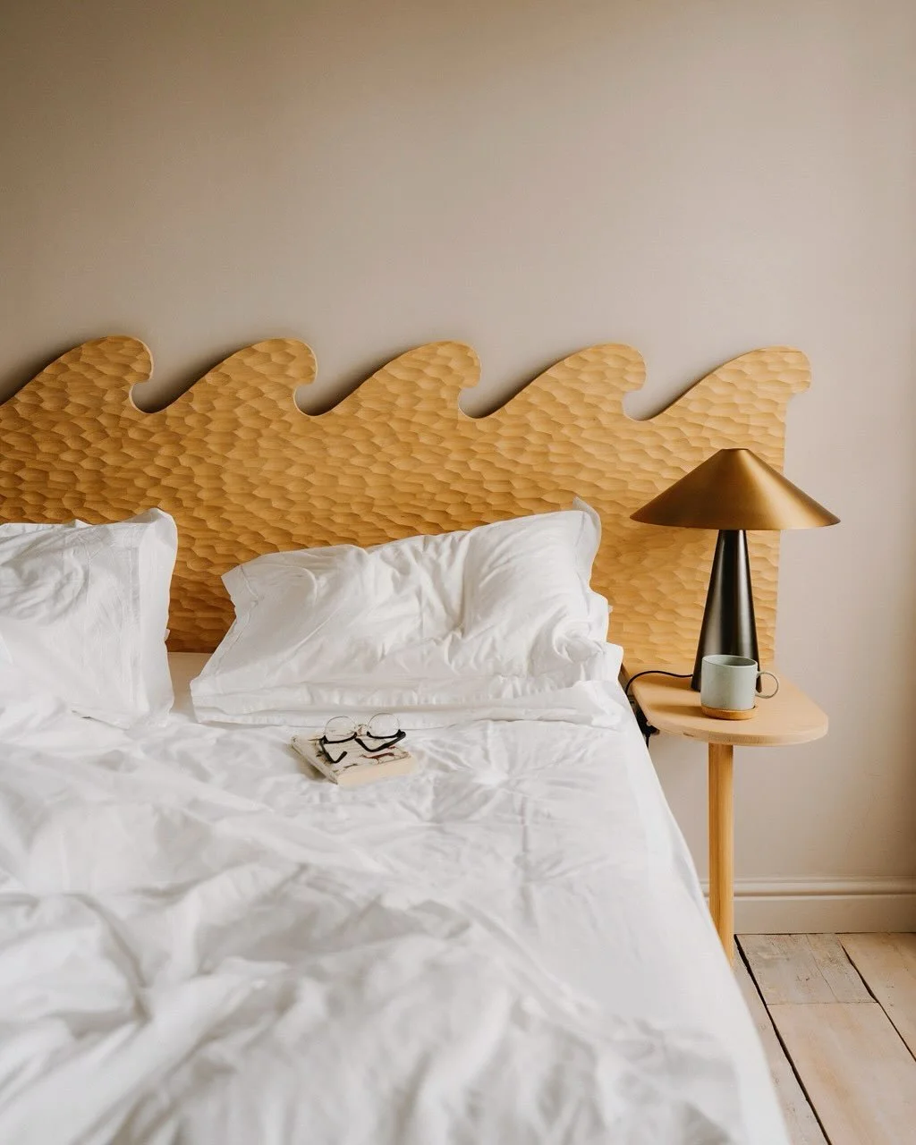 A neatly made bed with white sheets and pillows, with a pair of glasses and some books on the bed. There is a wooden nightstand with a modern table lamp and a mug, against a beige wall with a decorative wooden headboard.