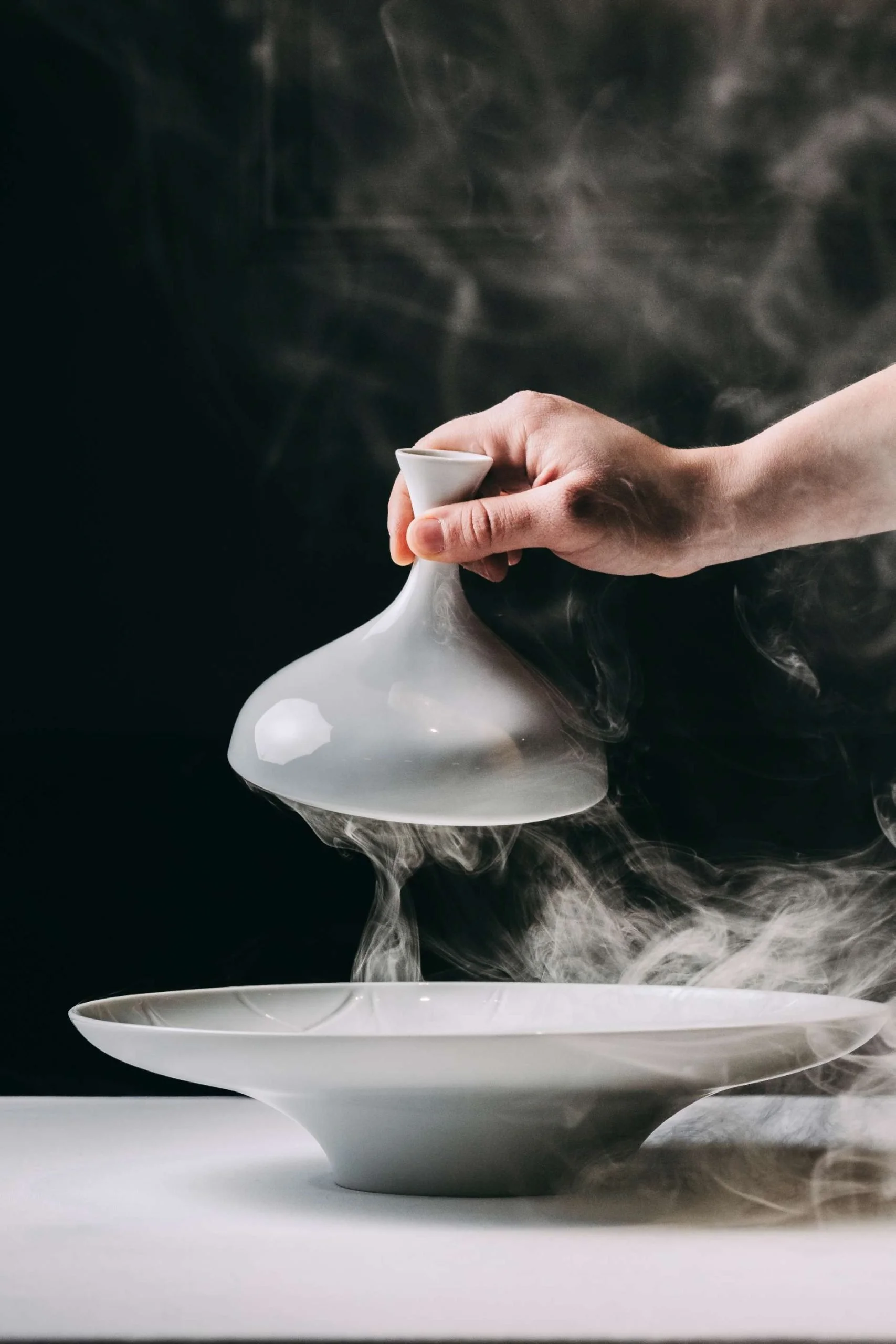 A person holding a small white ceramic vessel with steam rising from it, positioned above a matching ceramic bowl and tray, on a black background.