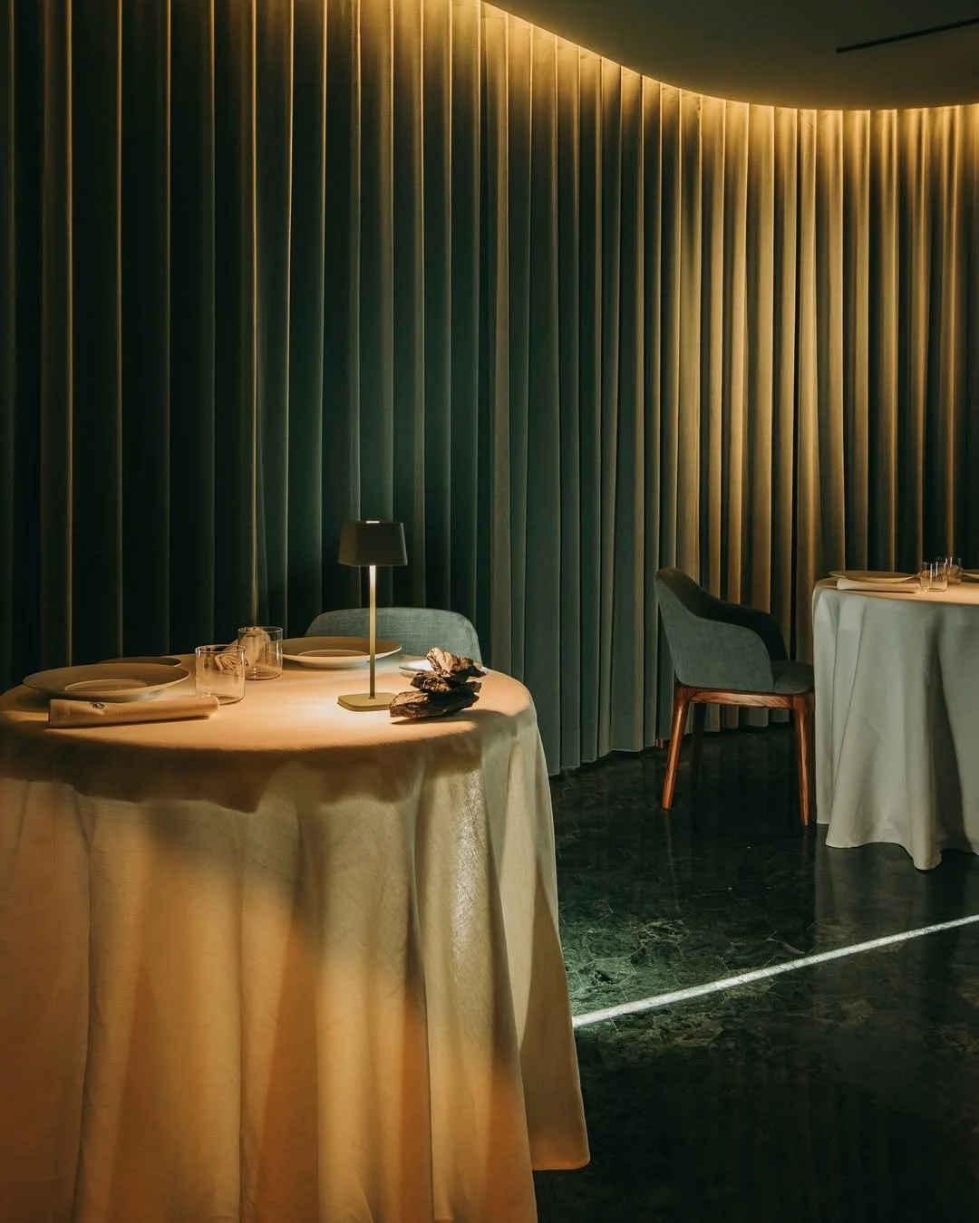 Empty restaurant with two round tables covered in beige tablecloths, set with plates, glasses, and napkins. A small lamp and decorative rocks are on one table. Dark green velvet curtains are in the background, and chairs are around the tables. Lighting is soft and warm.