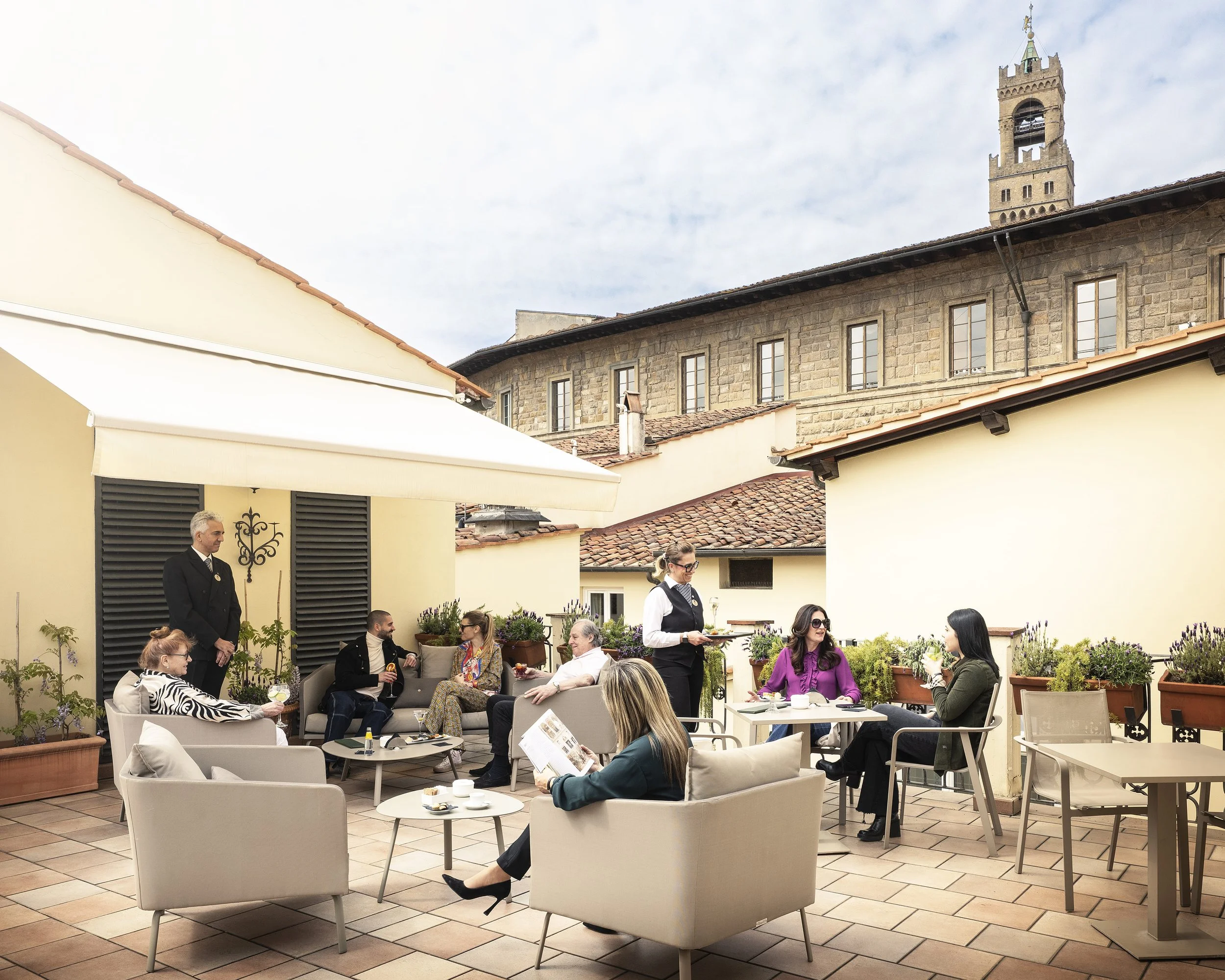 People dining and conversing on an outdoor terrace with chairs, tables, and potted plants, with a historic tower and building in the background under a cloudy sky.