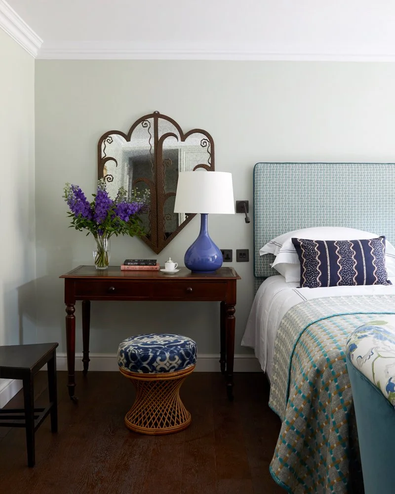 Bedroom corner with a wooden nightstand, a vase of purple flowers, a blue table lamp, a mirror on the wall, a bed with a blue headboard, and decorative pillows and bedding.