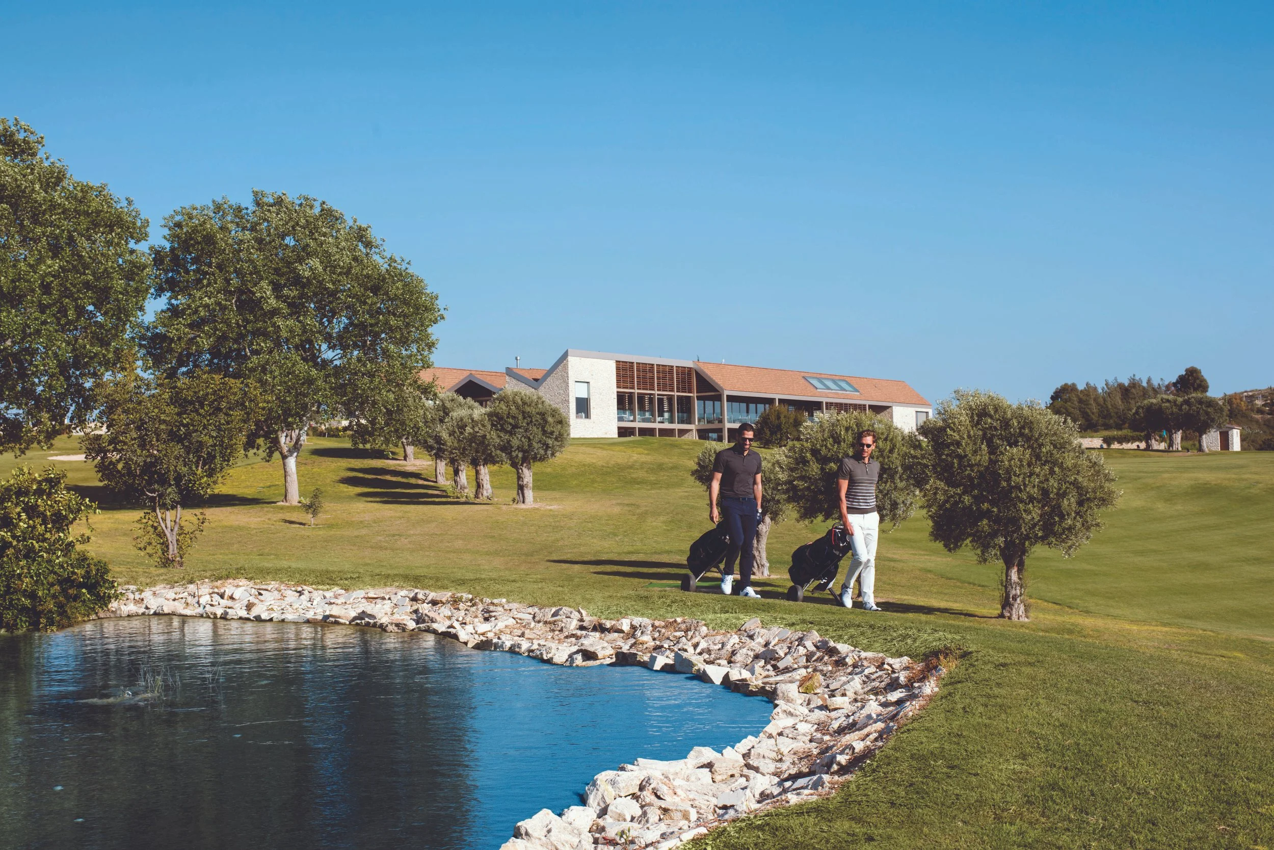 Two men walking along a grassy path near a pond, carrying luggage, with trees and a modern residential building in the background under a clear blue sky.