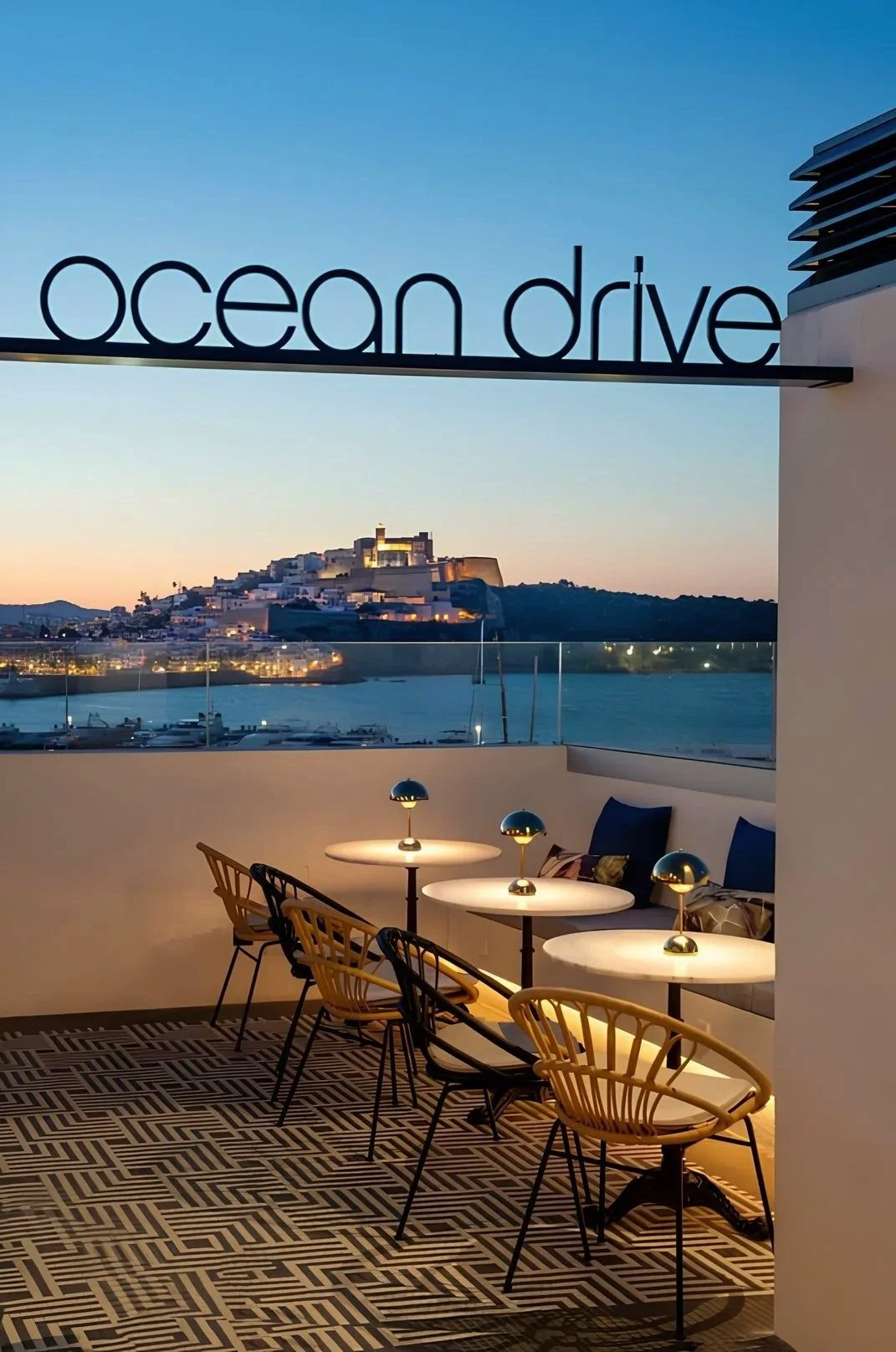 Outdoor dining area with four round tables and chairs, overlooking a marina with boats and a historic building on a hill during sunset. A sign with the words 'ocean drive' hangs above.