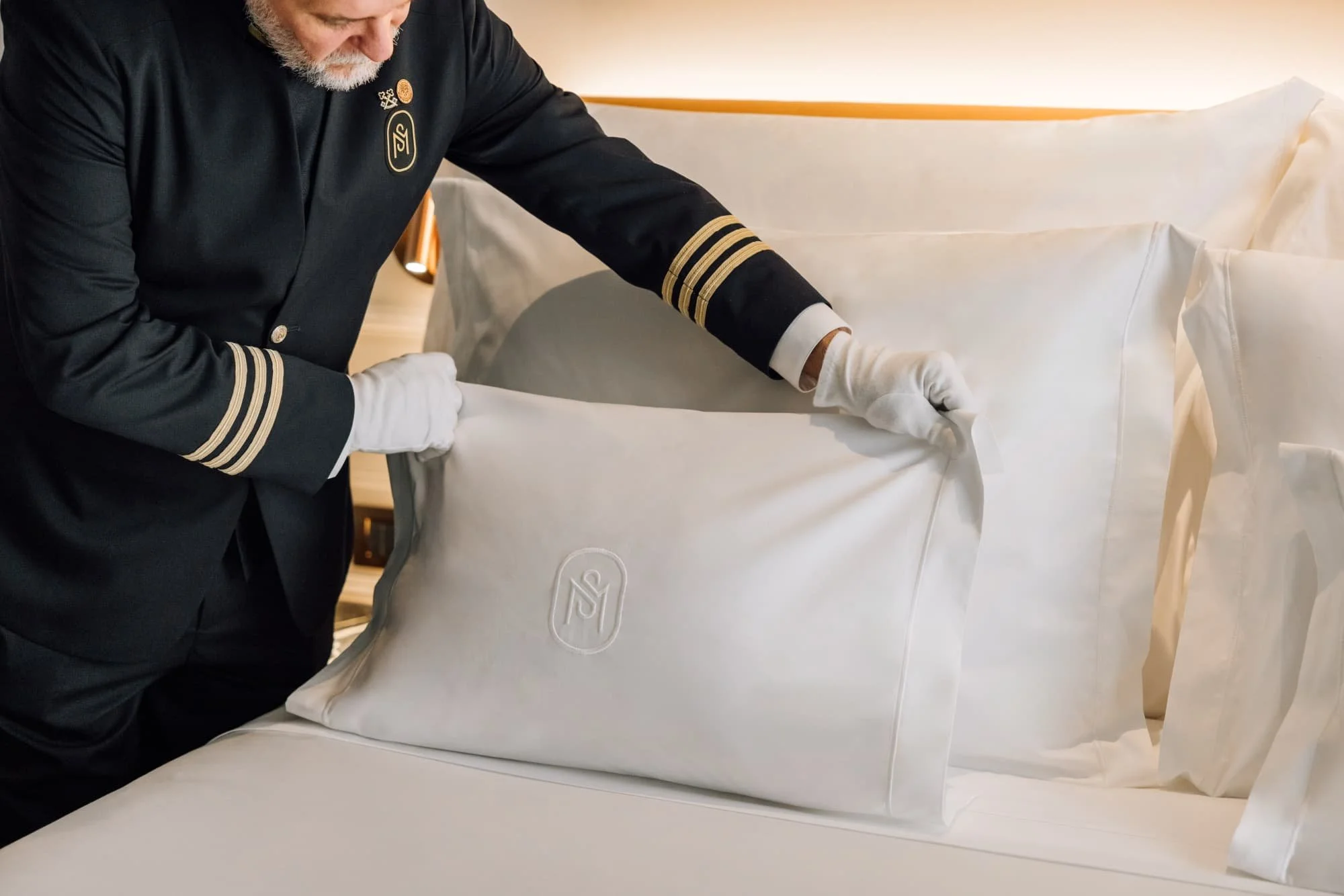 Person wearing a pilot's uniform making a bed with white sheets and a pillow in a hotel room.