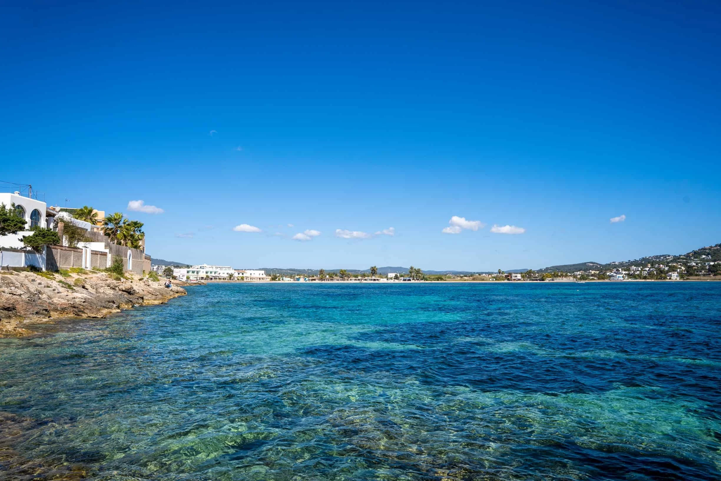 Clear blue sky over a coastal area with modern white houses, palm trees, and calm turquoise water in the foreground.