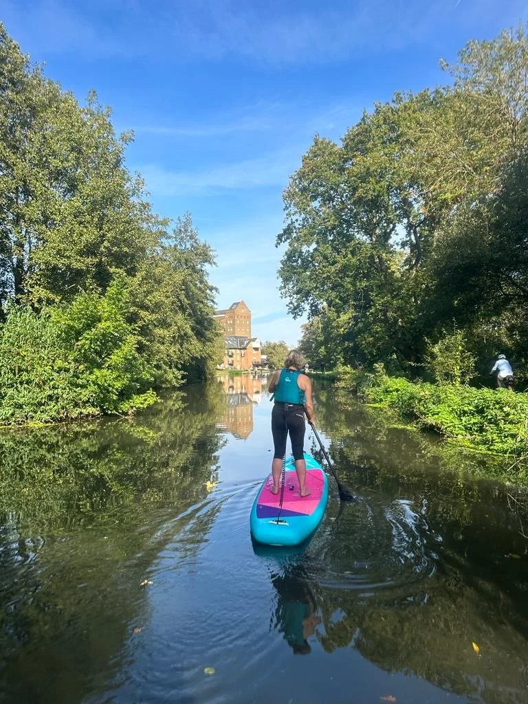 Person paddleboarding on a calm river surrounded by lush green trees under a clear blue sky.