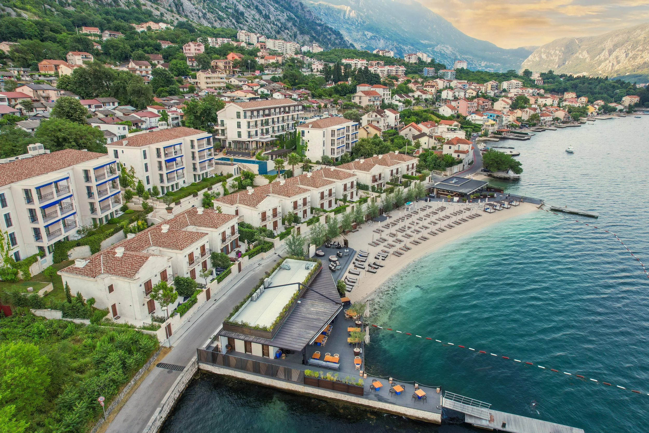 Aerial view of a coastal hillside town featuring modern white buildings with red-tiled roofs, a beach with lounge chairs, and a marina with boats along the water's edge, backed by lush green hills and mountains.