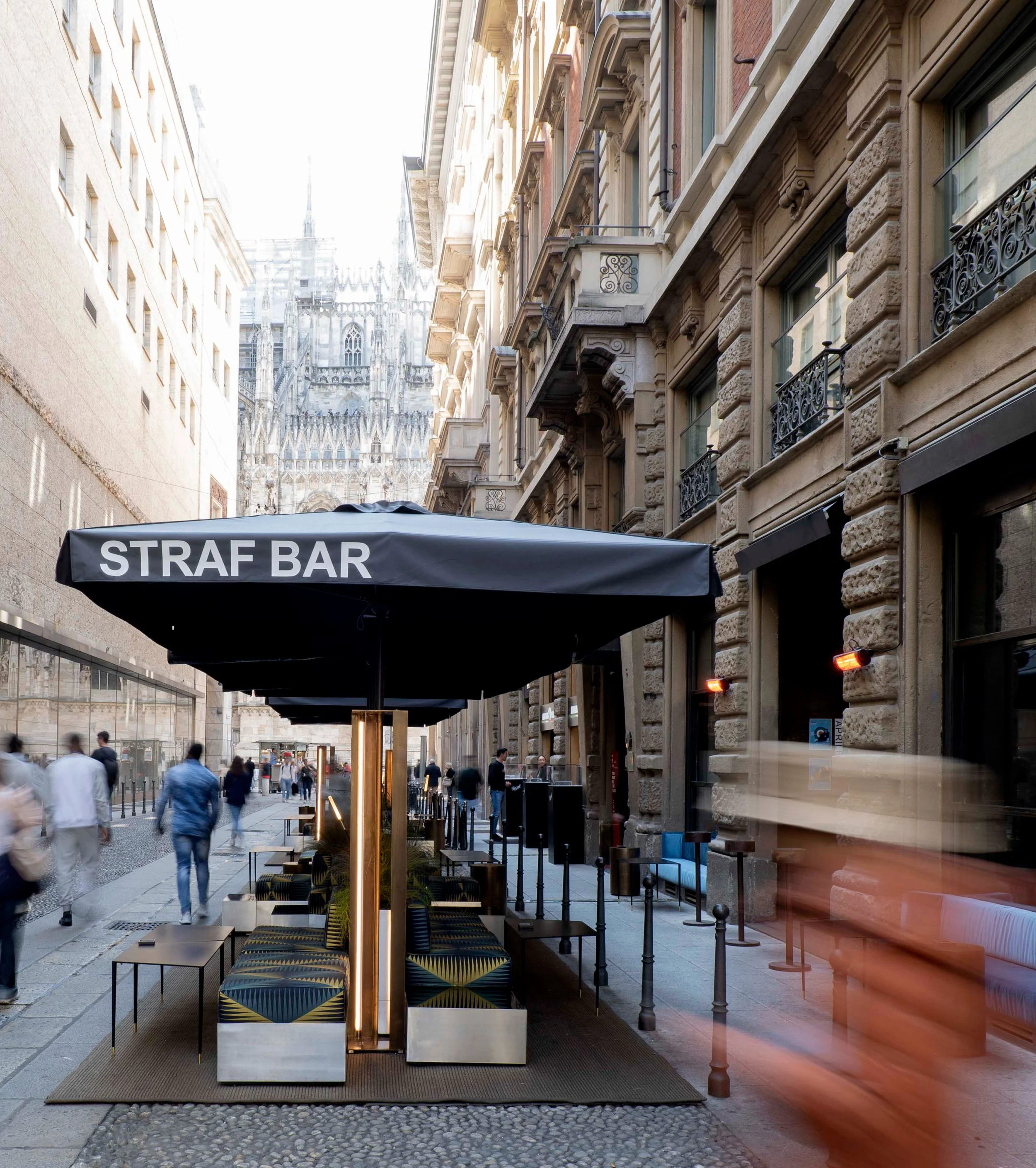 Outdoor seating area of a street bar with modern furniture and patterned cushions, located along historic European-style buildings with ornate architectural details, with the Cathedral of Milan in the background.
