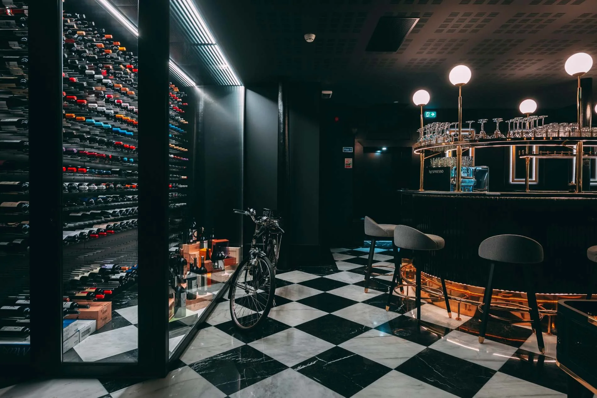 Interior of a modern bar or lounge with a wine display on the left, a bicycle near the center, and a circular bar counter with glassware and spherical lights above, all on a black-and-white checkered marble floor.