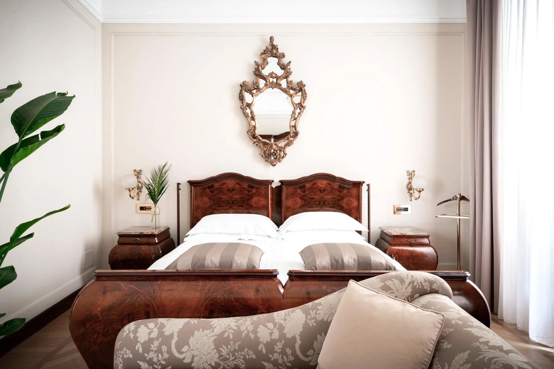 Elegant bedroom with two wooden beds, ornate mirror above, side tables, lamps, plants, and draped curtains.