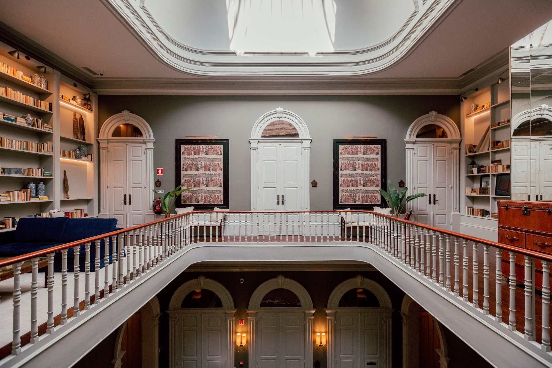 Interior of a multi-story building with a central open space, white railing, bookshelves on the sides, decorative framed artwork, green plants, and ornate architectural details.