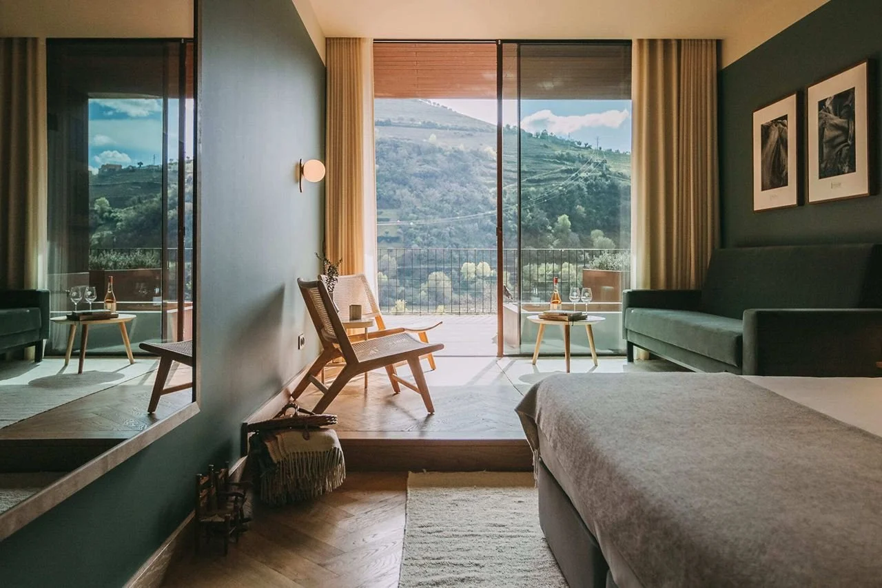 A hotel room with a large sliding glass door leading to a balcony, revealing a hillside landscape. The room has a bed in the foreground, a dark green sofa, two framed pictures on the wall, and wooden furniture including chairs and side tables with wine glasses.