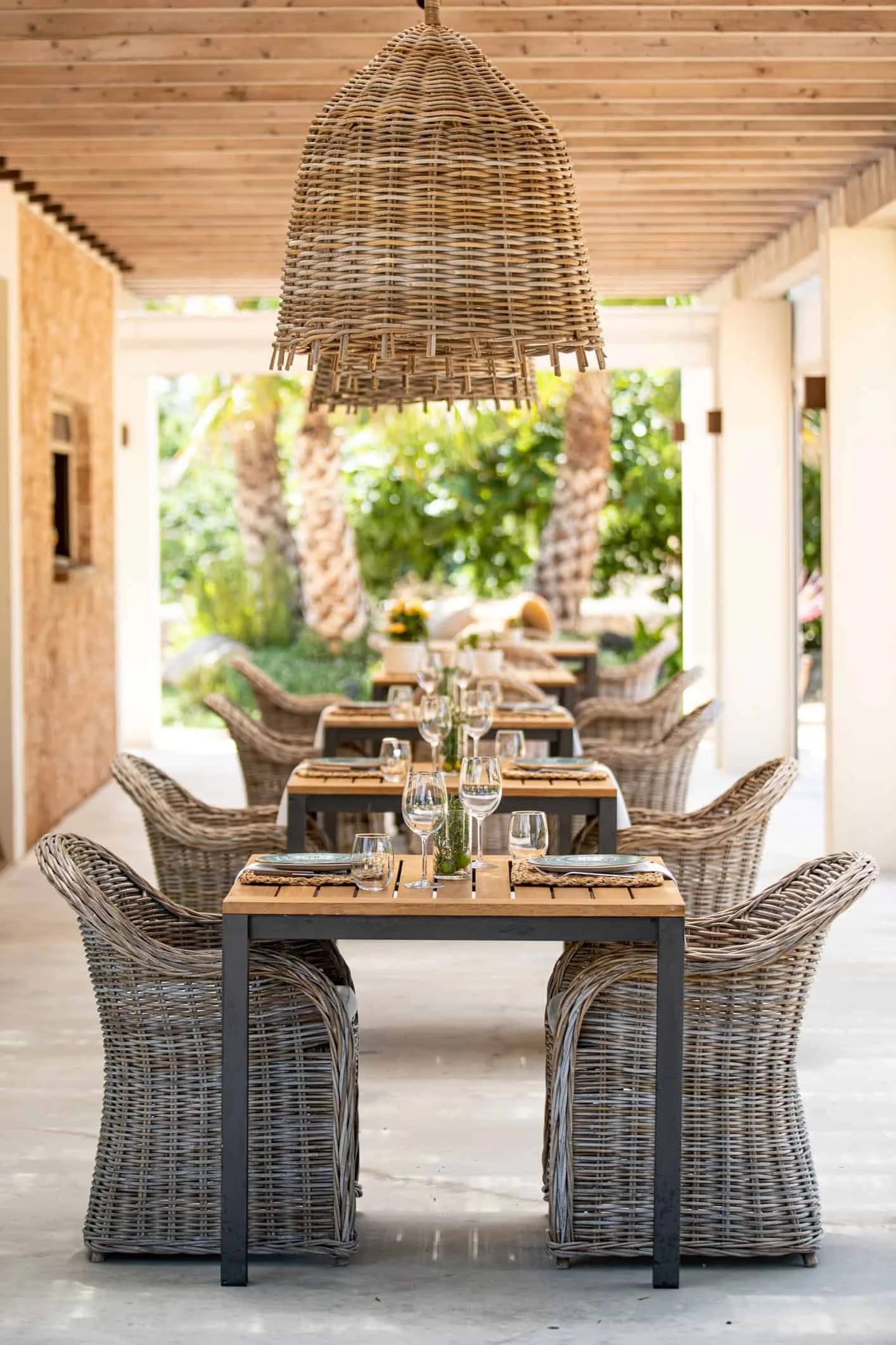 Table set for dining with chairs around it, and a woven rattan pendant light hanging above, in a bright outdoor or semi-outdoor space with greenery in the background.