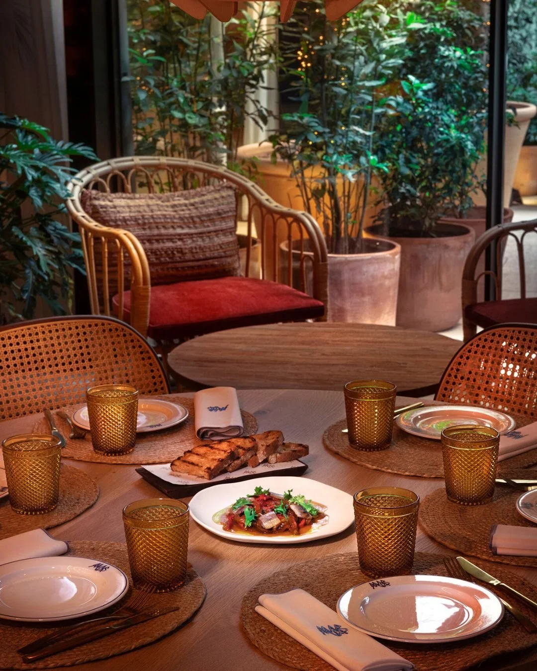 A dining table set for a meal with place settings, plates with food, and glasses, in a cozy restaurant with large potted plants and outdoor scenery visible through windows.