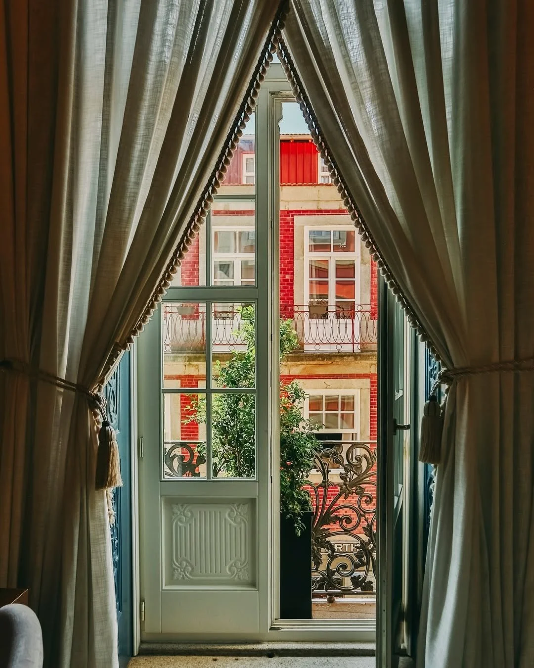 View from inside a room through a door and window, with curtains pulled to the sides, revealing a red brick building and potted plant on the balcony outside.