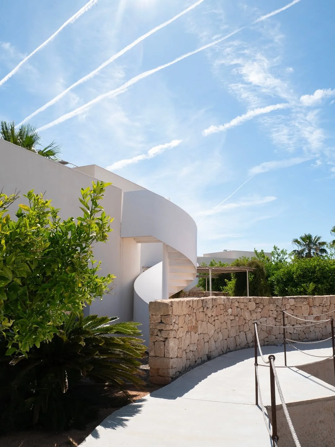 White modern building with an outdoor spiral staircase, stone wall, green plants, and a clear blue sky with streaks of white clouds.