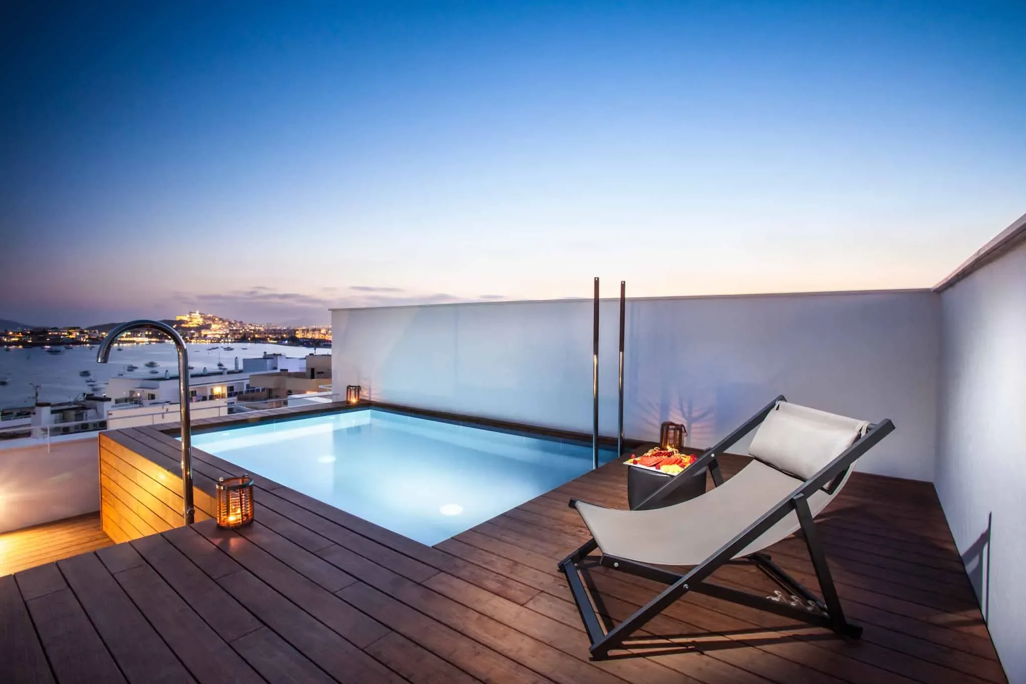 Rooftop swimming pool with lounge chair, picnic basket, and candles during dusk, overlooking city skyline.