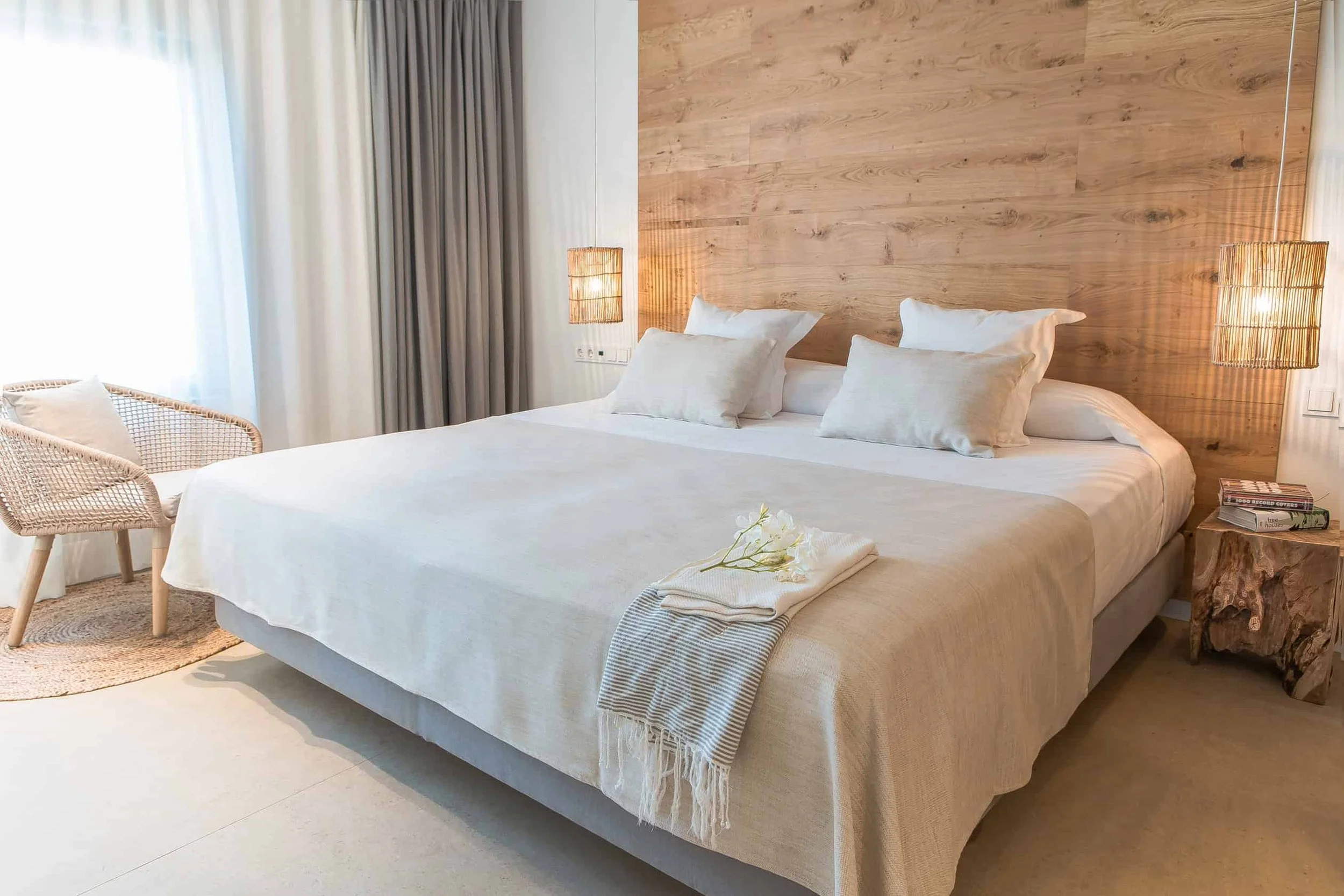 A neatly made bed with white linens and pillows, a wooden headboard, a wicker chair, and a nightstand with books and a decorative object, in a room with light-colored flooring and a mix of white and dark gray curtains.