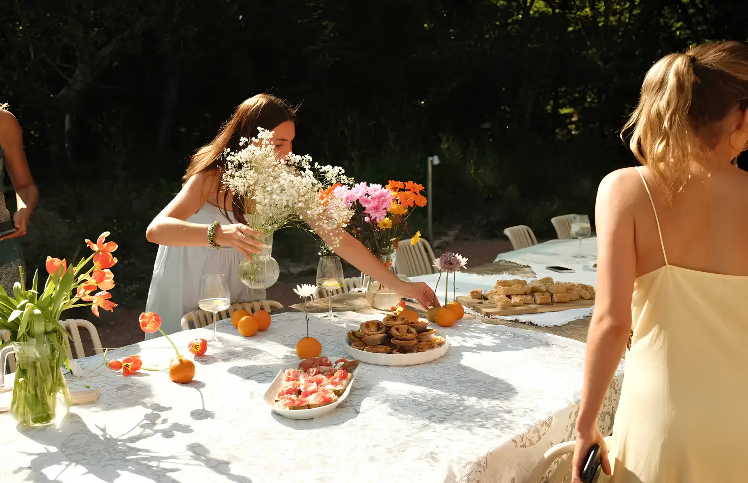 Outdoor celebration with a long table decorated with flowers, food, and drinks, surrounded by women and trees.