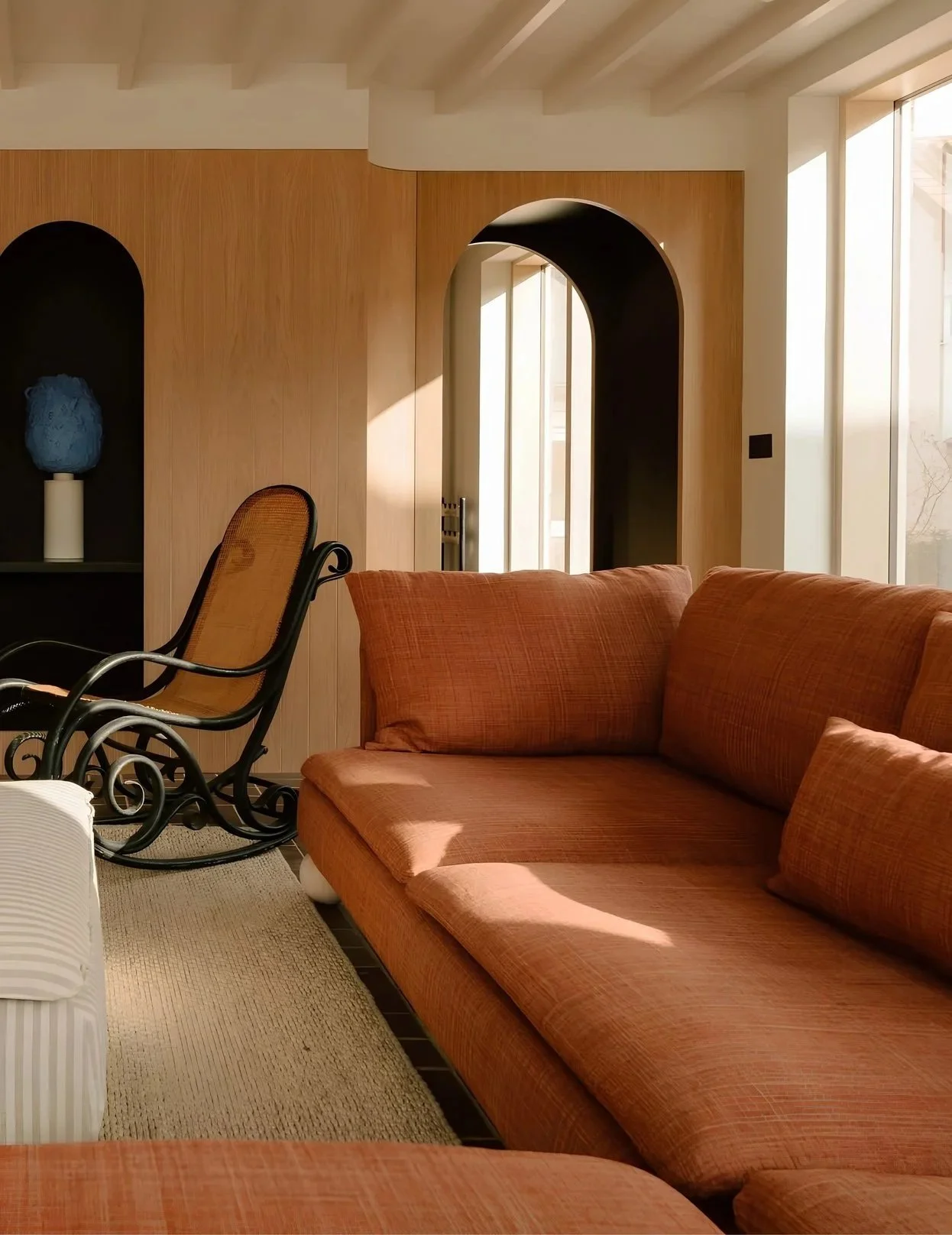 Living room with orange sofa, black rocking chair, wooden wall, and large windows letting in natural light.