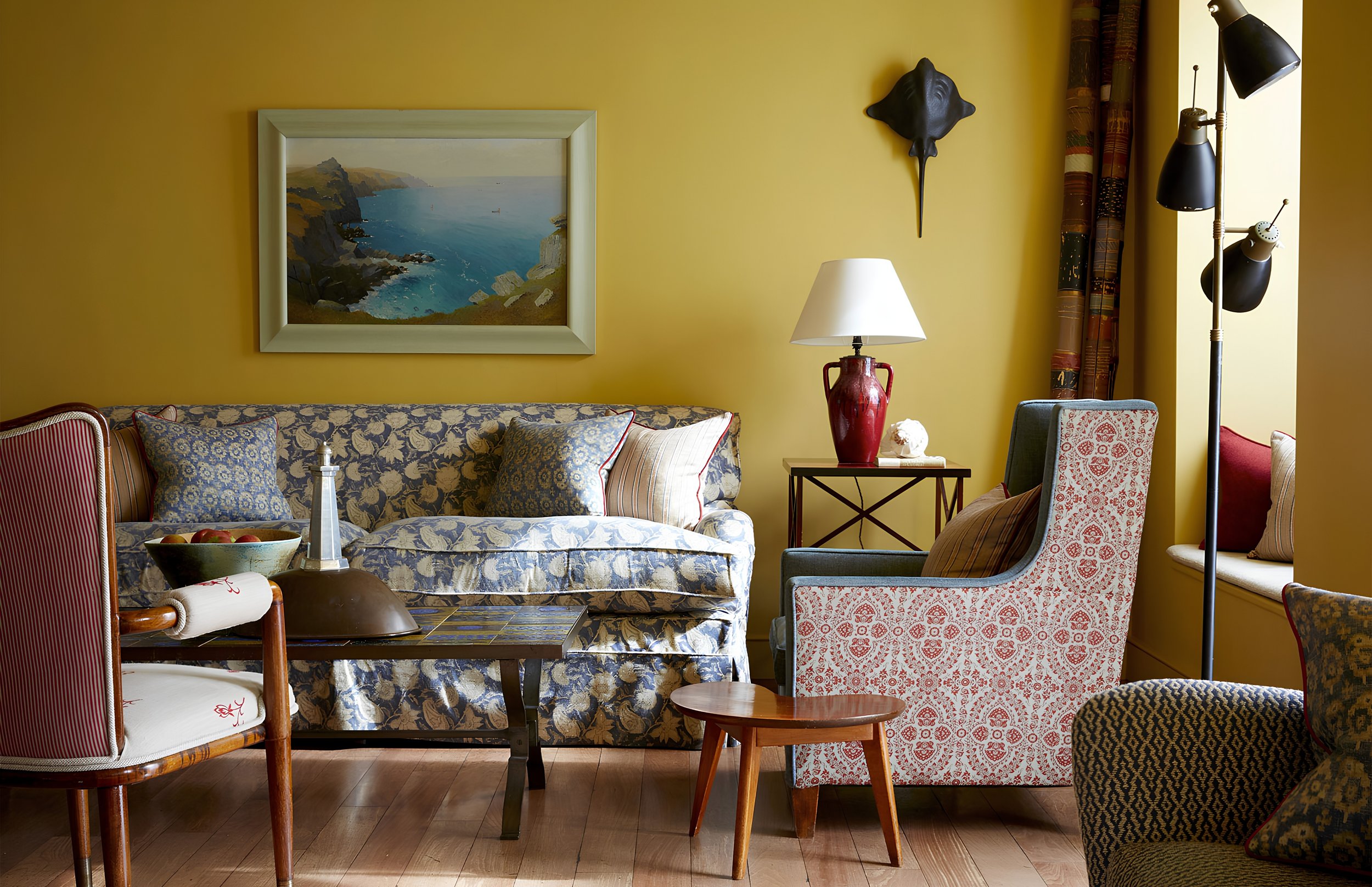 A cozy living room with a yellow wall, featuring a framed landscape painting, a dark wall mask, a white-and-red patterned armchair, a blue floral sofa with pillows, and a side table with a red vase lamp, a smaller white rose sculpture, and a chair wi