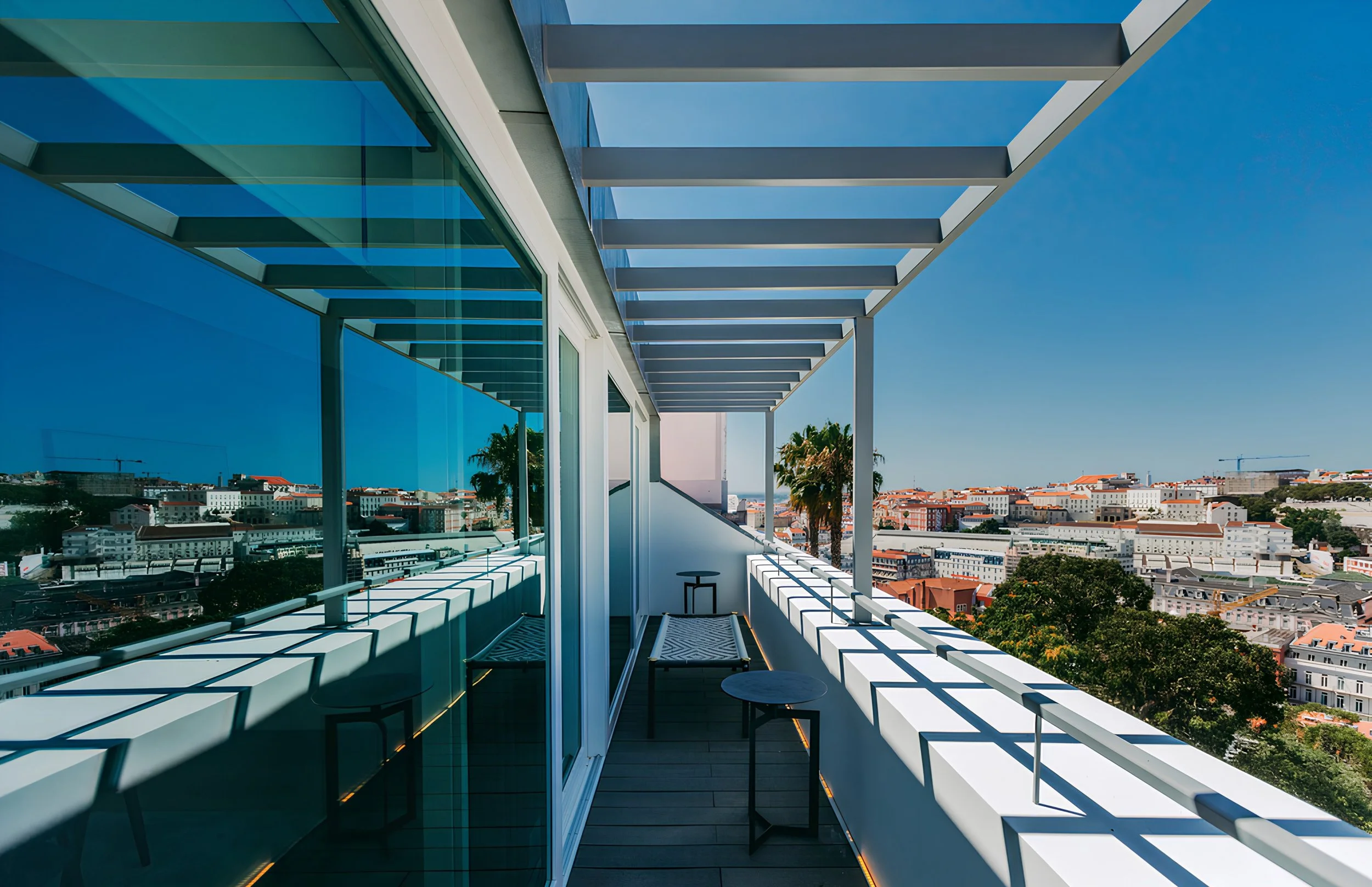 Balcony with modern white railings and benches, overlooking a cityscape with buildings and trees under a clear blue sky.