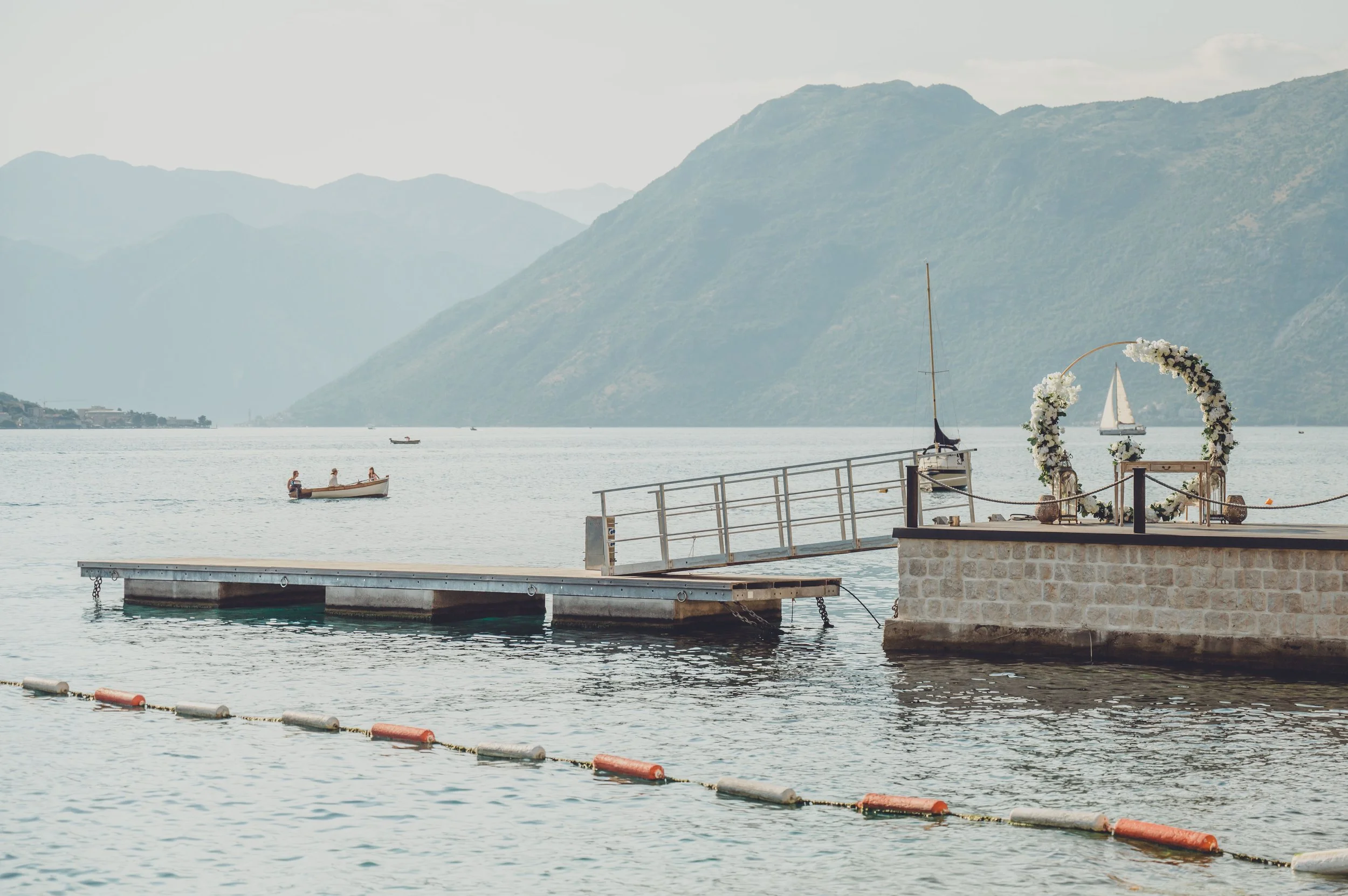 A lakeside scene featuring a small dock with a decorative archway decorated with white flowers, a ladder leading to the water, and several boats including a sailboat, with mountains in the background under a partly cloudy sky.