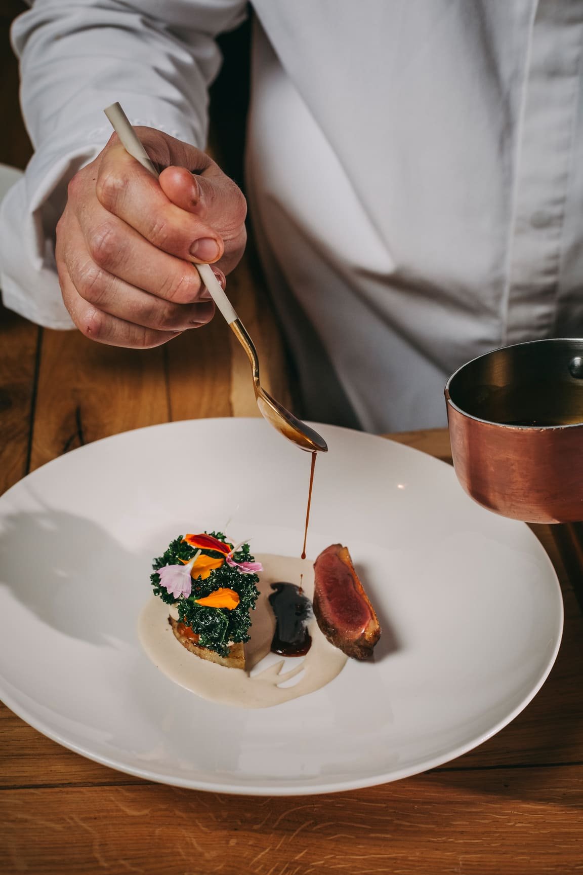 Close-up of a chef in a white uniform drizzling sauce over a gourmet dish on a white plate, featuring a piece of seared meat, vegetables, and garnishes, with a copper sauce pot nearby.