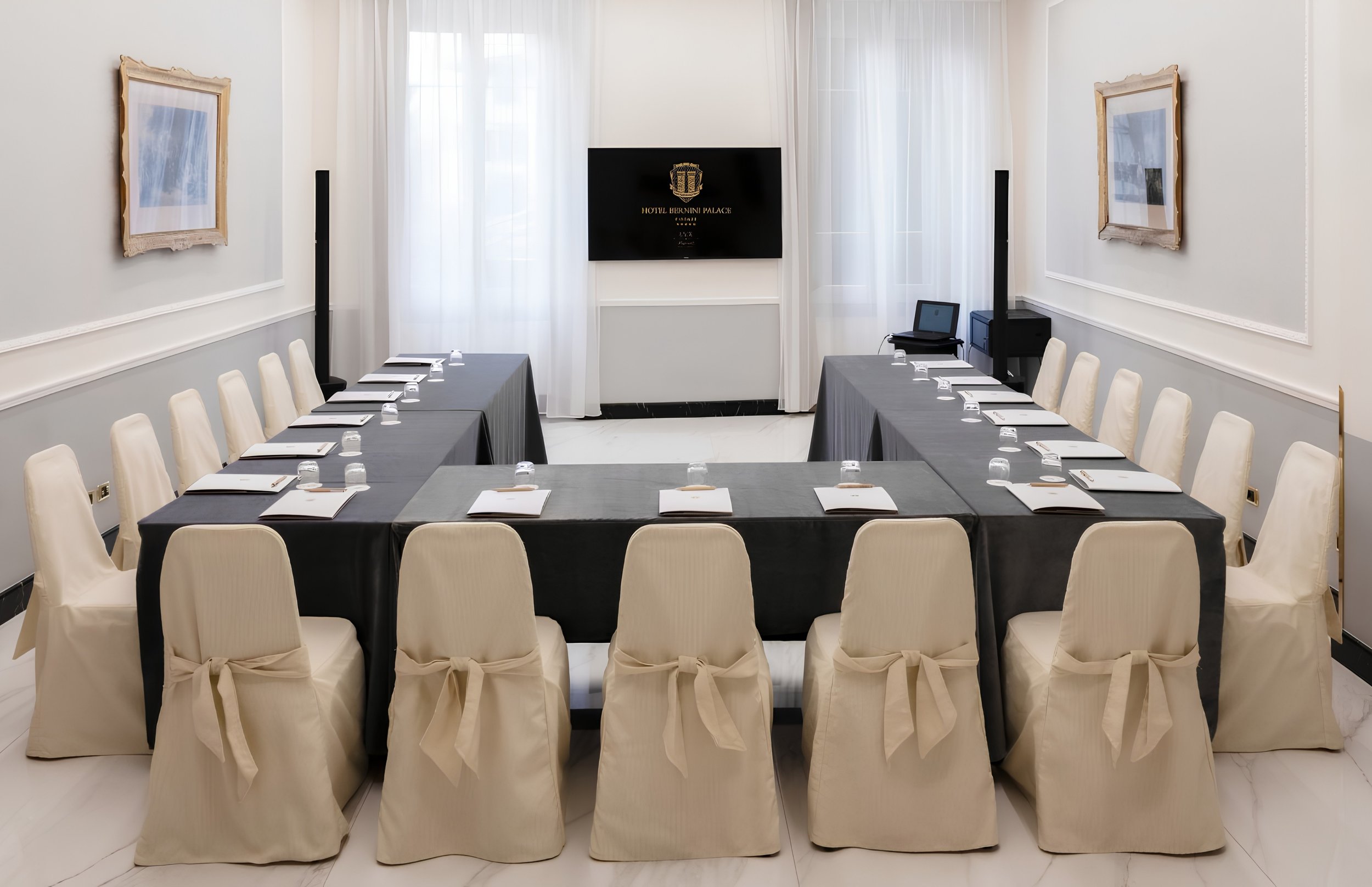 A conference room with a U-shaped arrangement of tables covered in dark tablecloths, each set with notepads, pens, and water glasses. Cream colored chairs with bows on the back surround the tables. The room has white walls with two framed paintings and a large flat-screen TV displaying the logo of Hotel Brunini Palace.