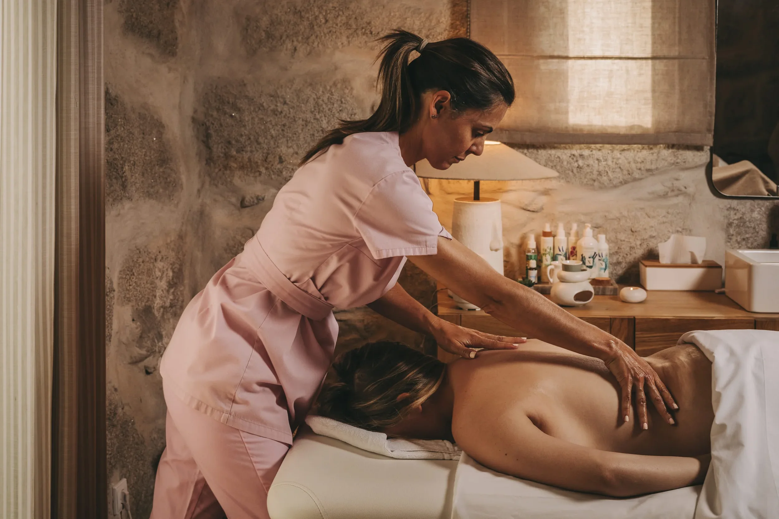 Massage therapist giving a massage to a client lying face down on a massage table in a spa room with warm-colored walls and various massage oils and supplies on a wooden shelf.